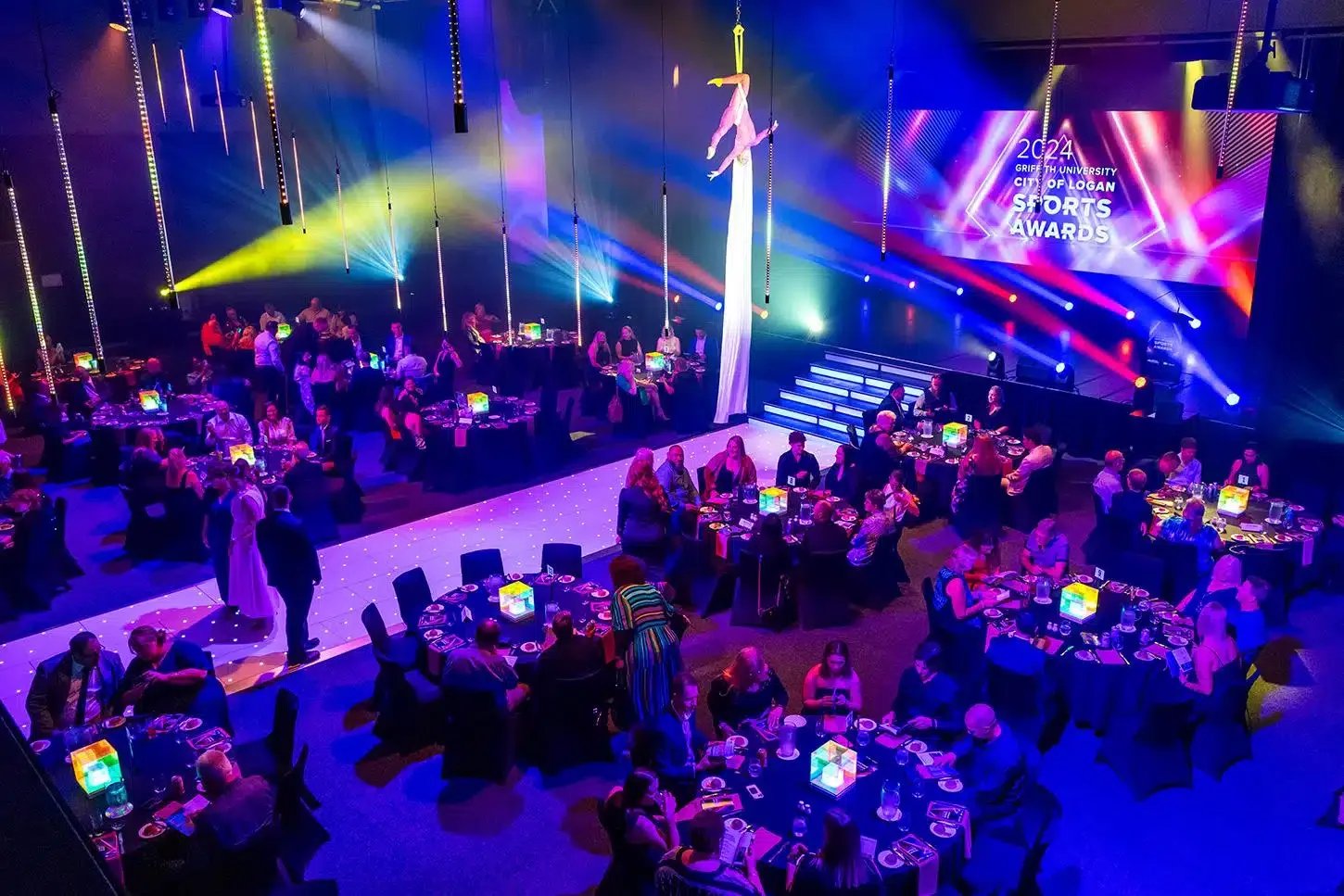A large event hall set up for an awards ceremony with multiple dining tables, colorful stage lighting, a stage with a screen displaying '2024 Griffin University City of Logan Sports Awards,' and aerial acrobats performing on fabric in the center of the room.