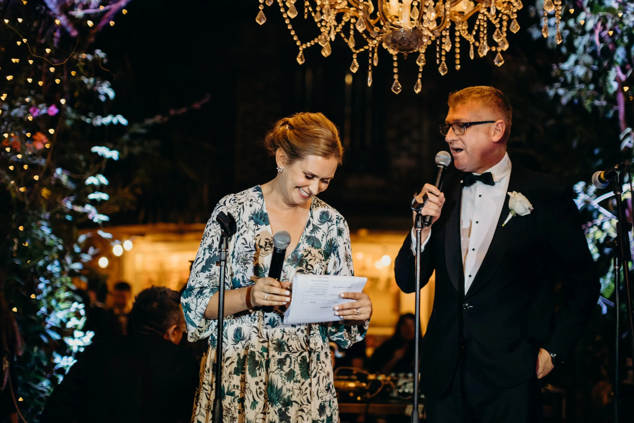 A woman in a floral dress reads from notes while smiling, and a man in a tuxedo with glasses and a white boutonniere speaks into a microphone at a wedding reception with a chandelier and guests in the background.