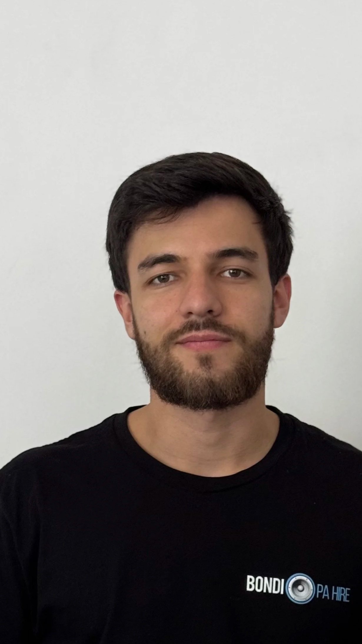 A young man with short dark hair and a beard, wearing a black T-shirt with a logo that says 'BONDI PA HIRE' and a speaker icon, standing in front of a plain white wall.