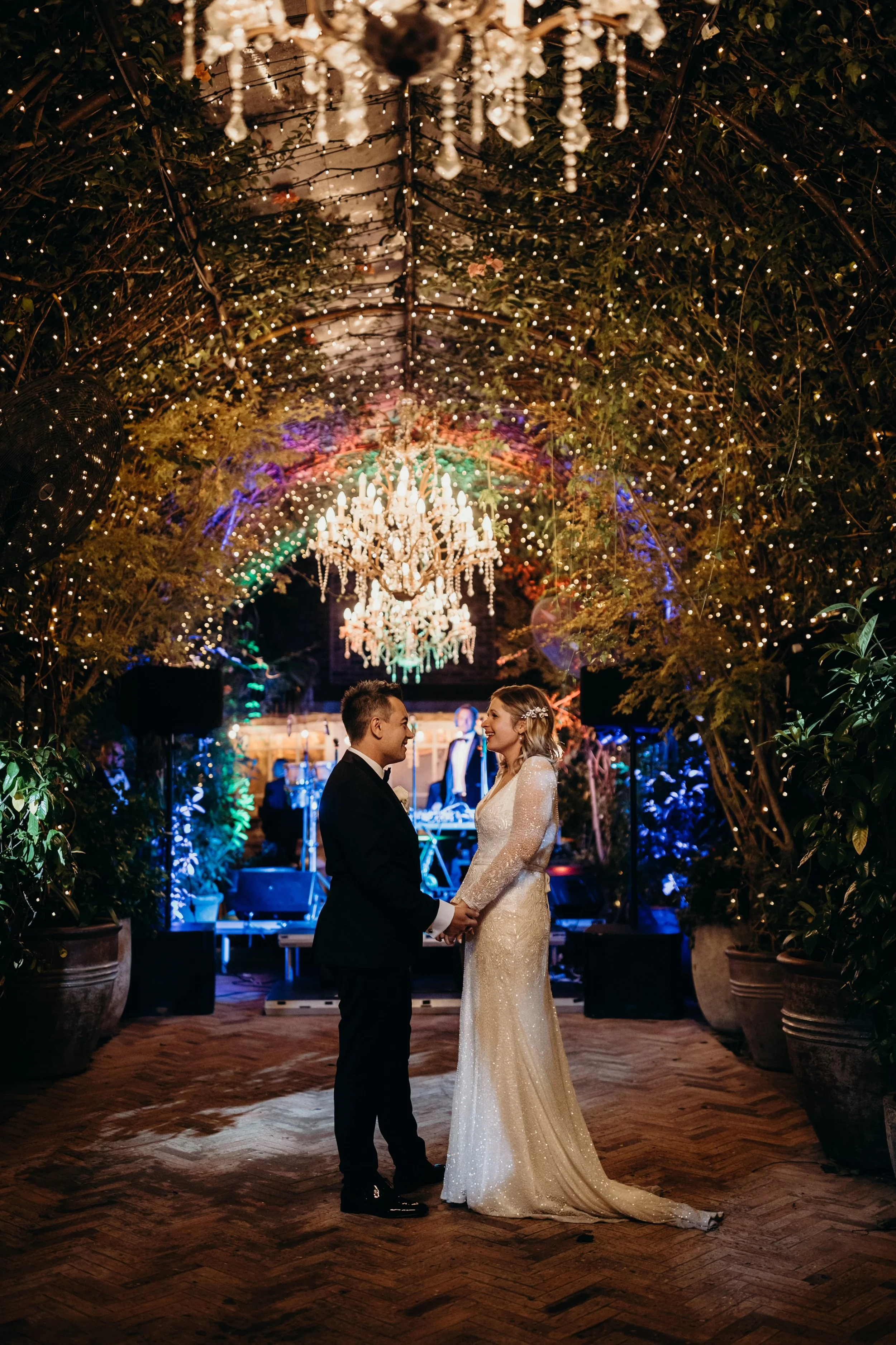 A bride and groom holding hands and facing each other during their wedding ceremony in an elegant, decorated venue. The ceiling is adorned with strings of lights and a grand chandelier, creating a romantic ambiance.