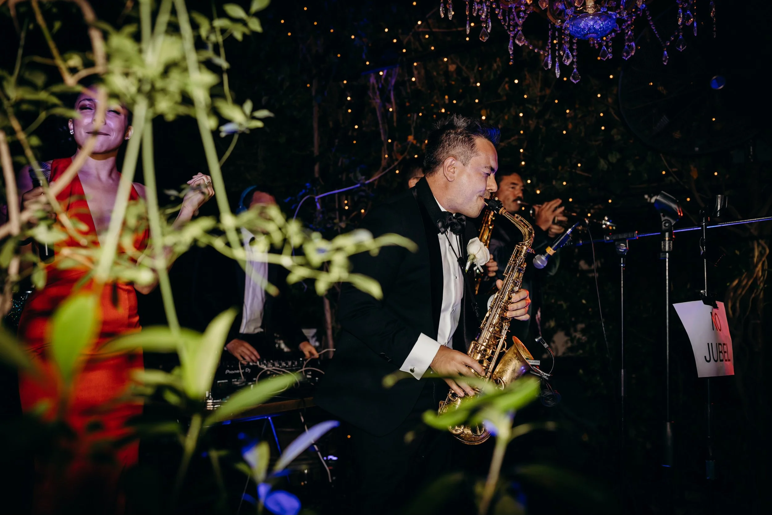 Musicians performing at an indoor event, with one man playing a saxophone and others singing or playing instruments, under decorative lighting and chandeliers, with a sign reading 'No JUBEL' in the background.
