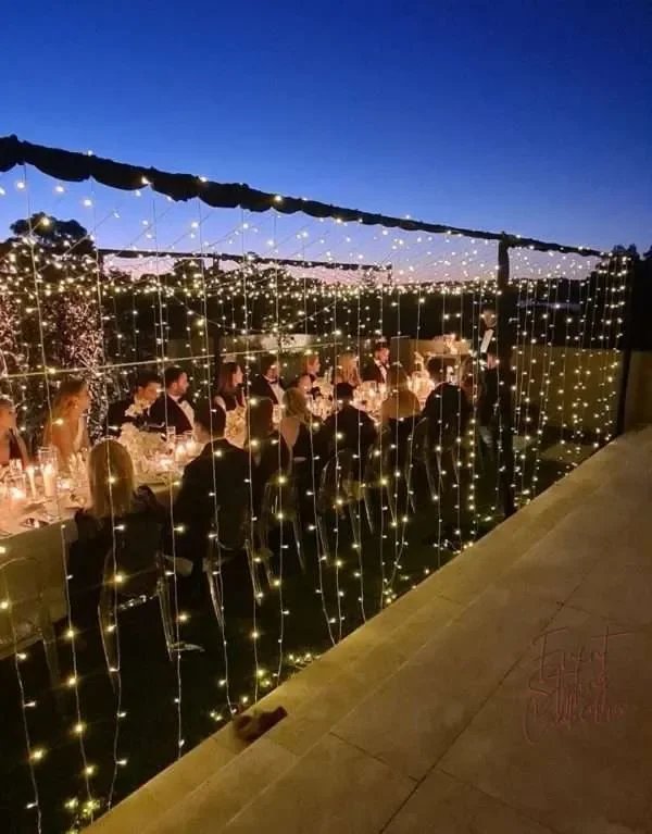 An outdoor evening dinner party with a long table decorated with string lights hanging from a canopy, illuminated by candles, with several guests seated and a dark blue sky in the background.