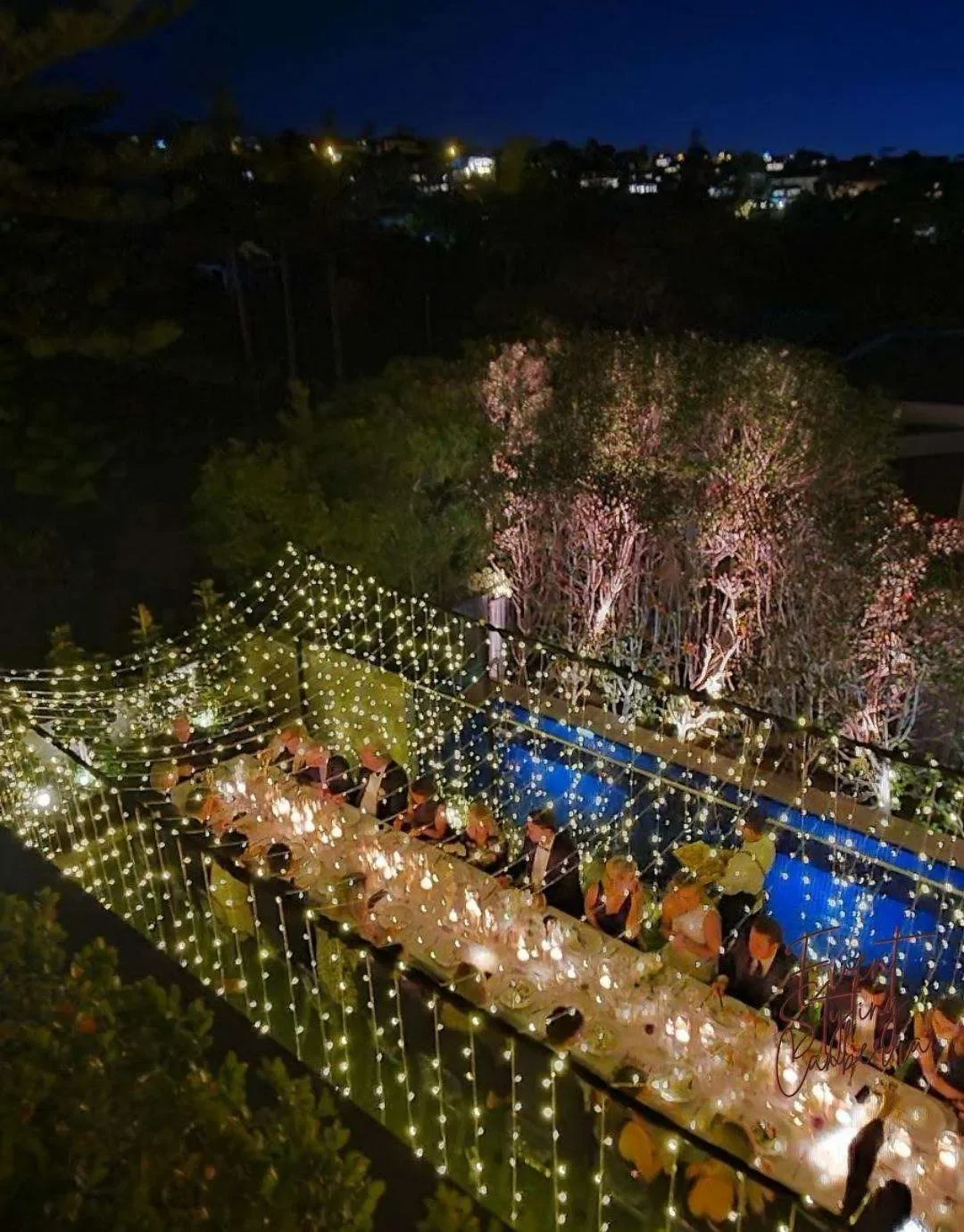 Overhead view of a nighttime celebration with a long banquet table decorated with string lights, surrounded by trees with pink blossoms, set outdoors on a terrace or garden area.
