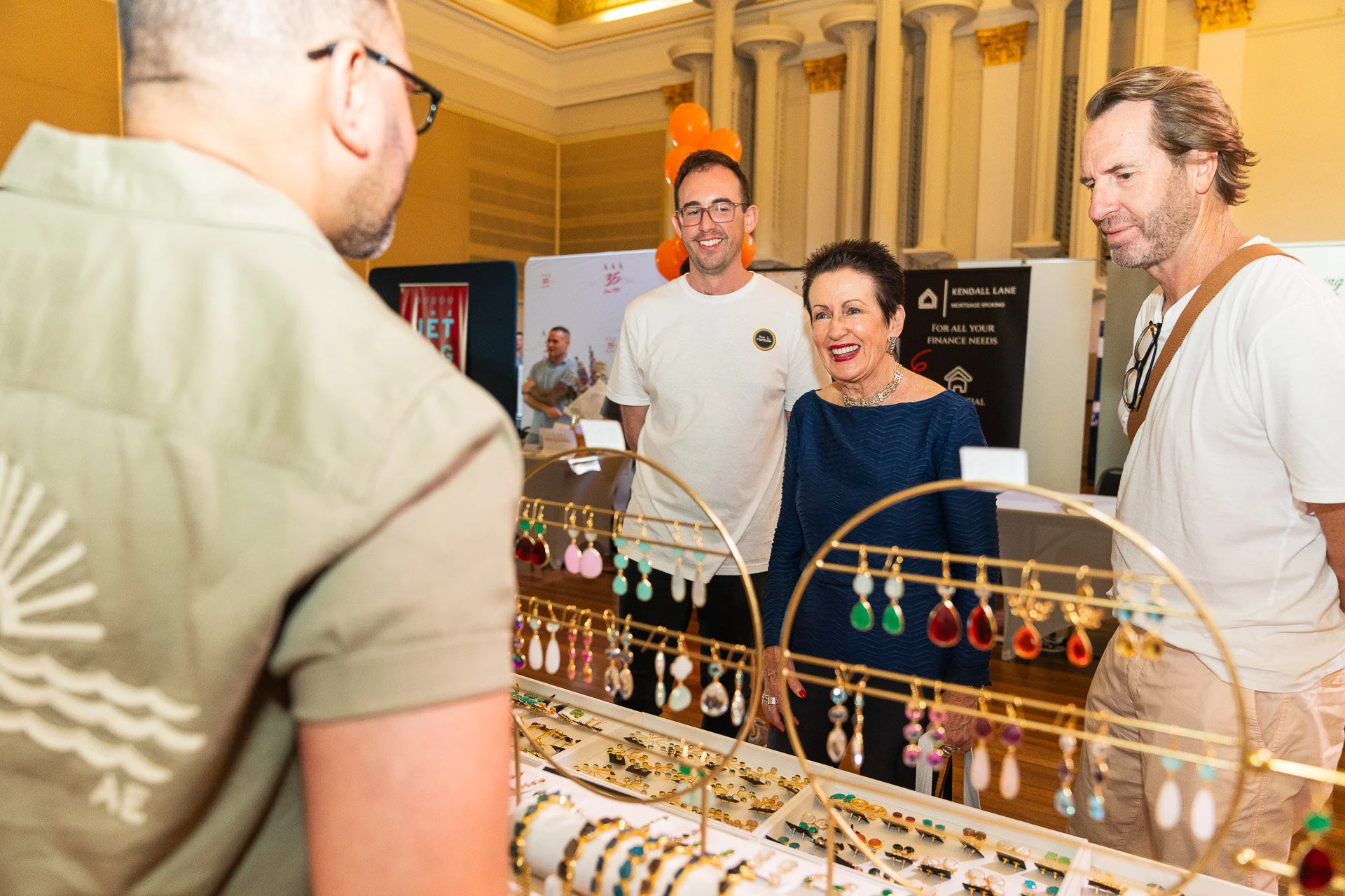 People shopping for jewelry at a trade show or market, with jewelry displays and vendor booths in the background.