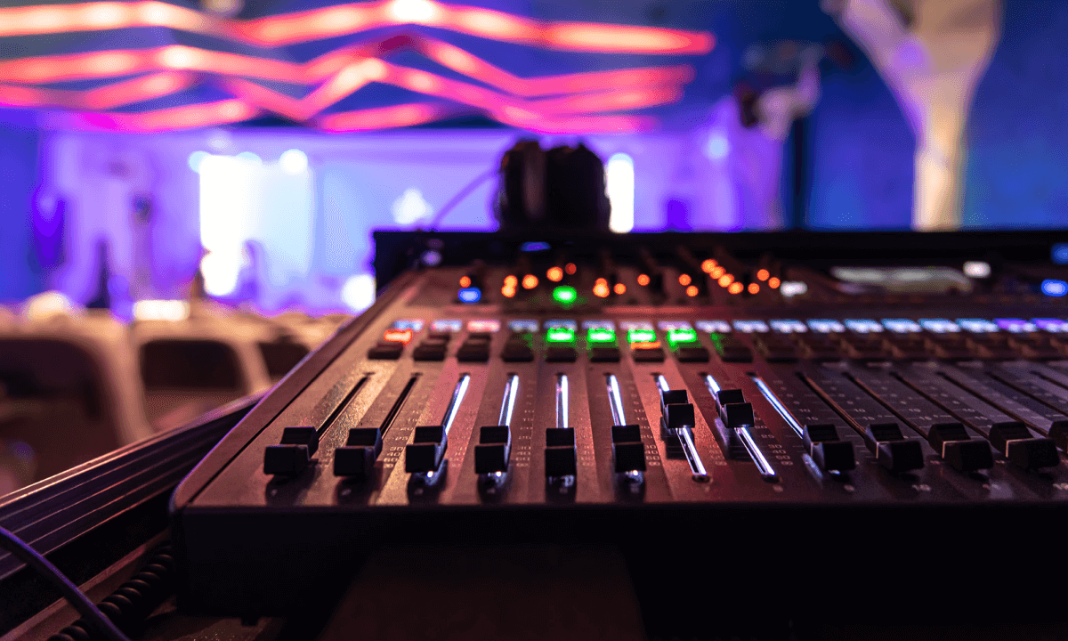 Close-up of a professional audio mixing console with colorful lights in a dimly lit venue or event space.