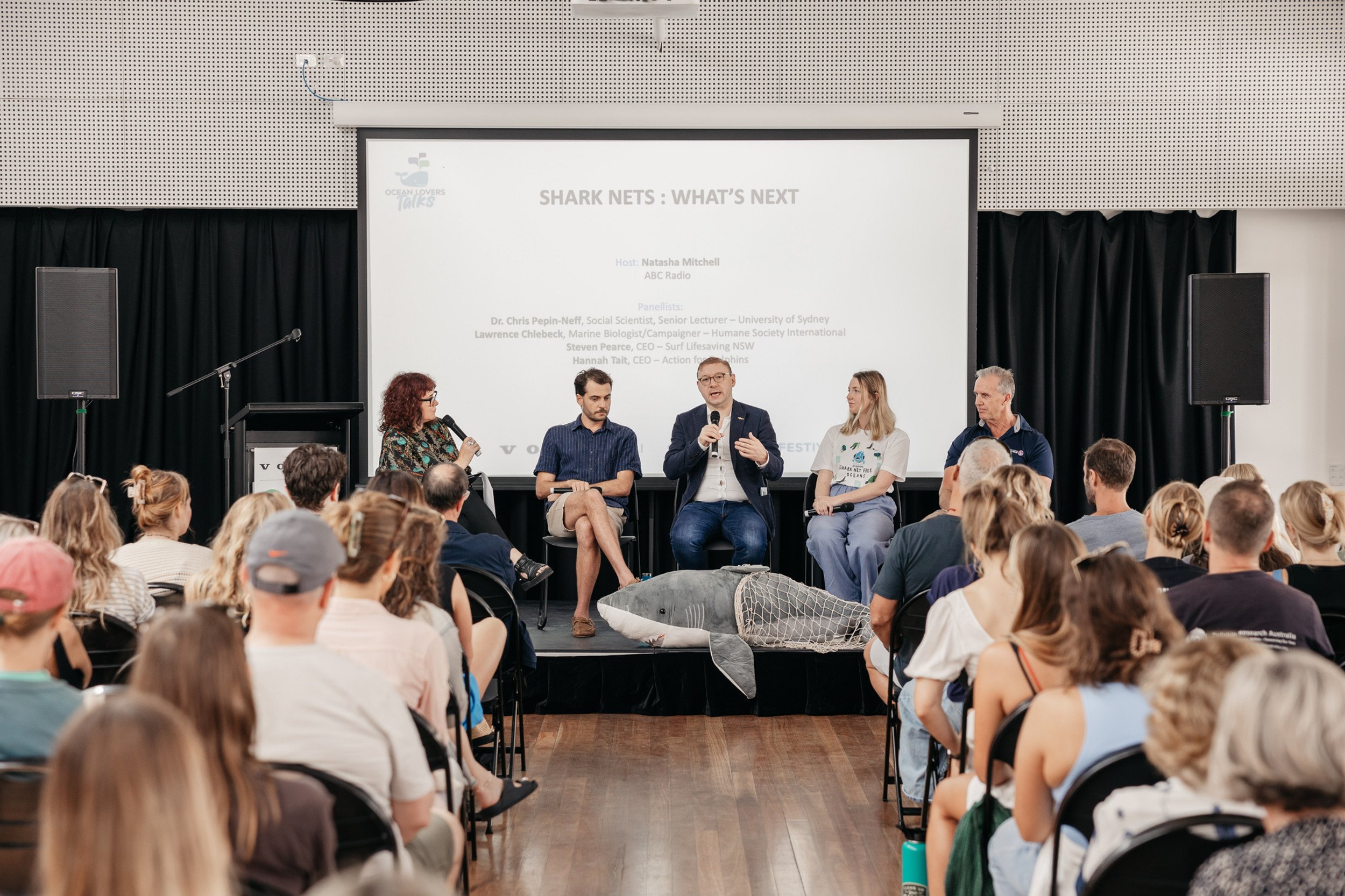 Panel discussion at a conference with five people on stage, one holding a microphone, with an audience watching. There is a large screen behind them displaying the event title and panelists. A large inflatable shark is on the stage floor in front of the panel.