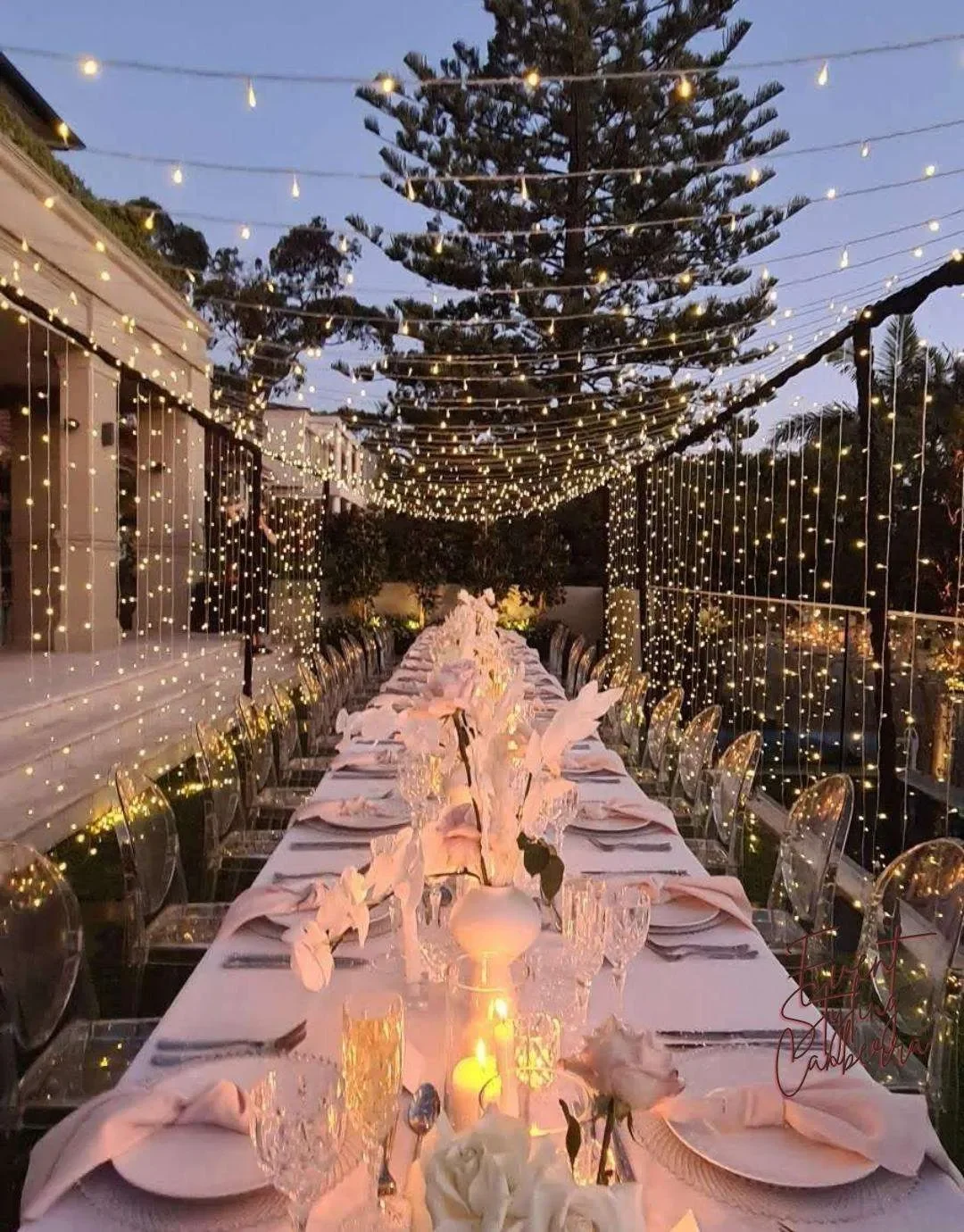 An elegantly decorated outdoor dining table illuminated by string lights, with white floral centerpieces, candles, glassware, and neatly folded pink napkins set for a celebration during dusk.