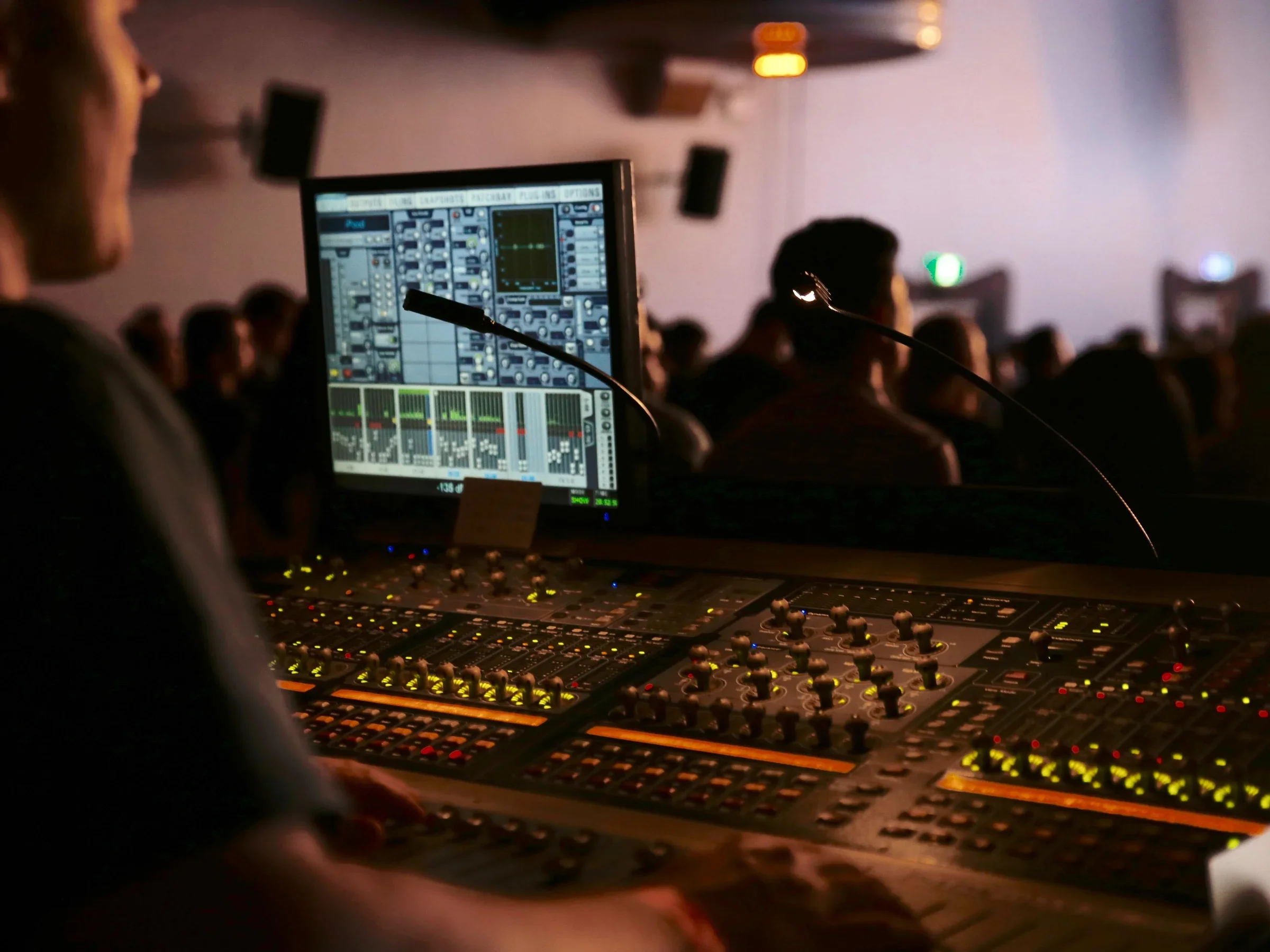 Close-up of a sound engineer operating a professional audio mixing console at a live event with an audience in the background.