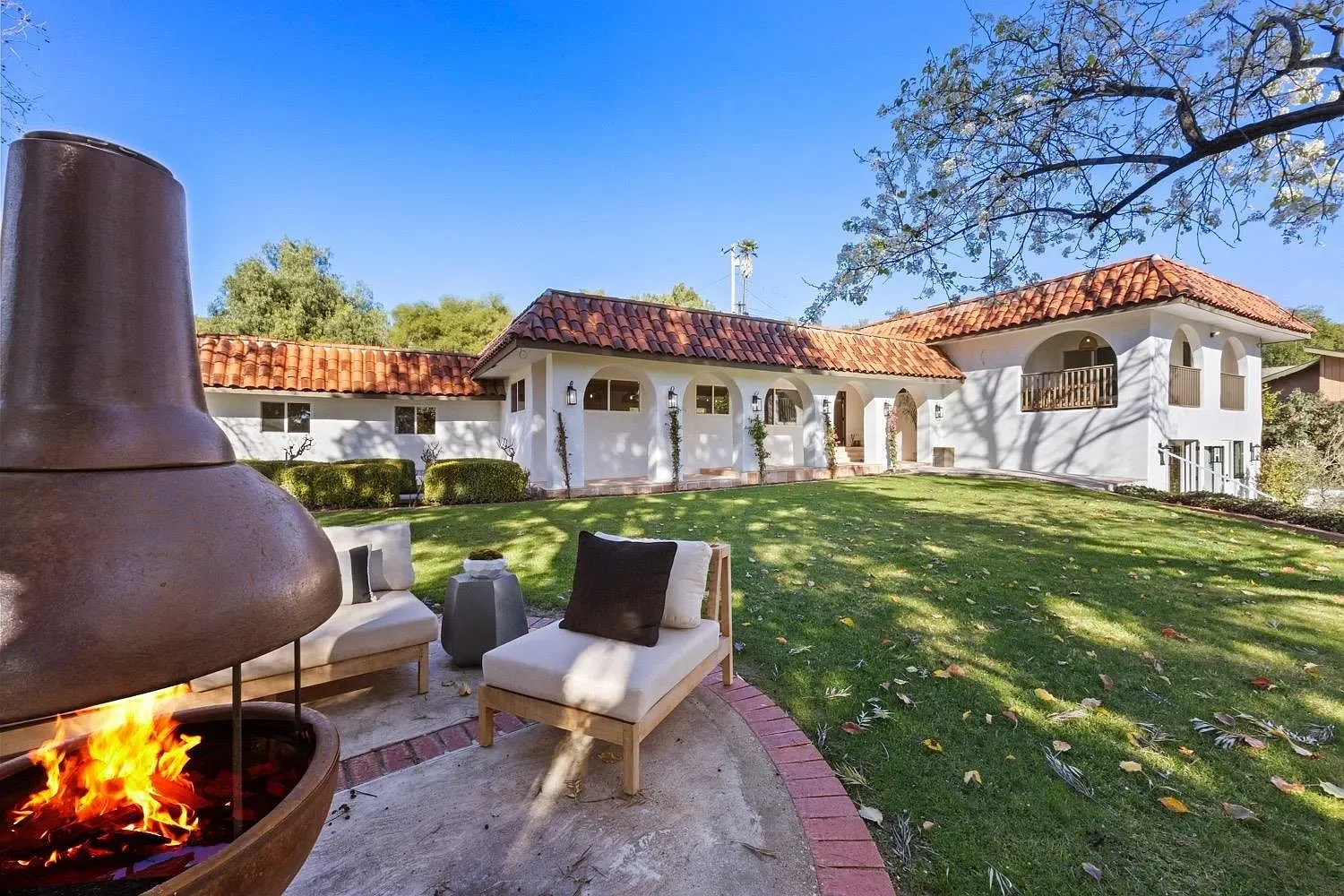 A backyard scene with a white house, red tiled roof, and small balcony. There is a grassy lawn with shadows from trees, a fire pit, a cushioned outdoor chair, and other patio furniture.