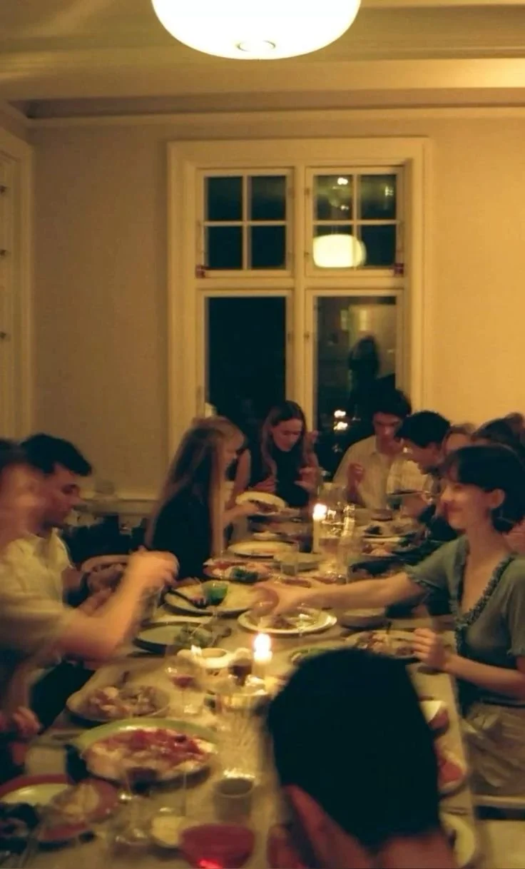 Group of people sitting around a dining table with pizza and drinks, in a warmly lit room with beige walls and a window in the background.