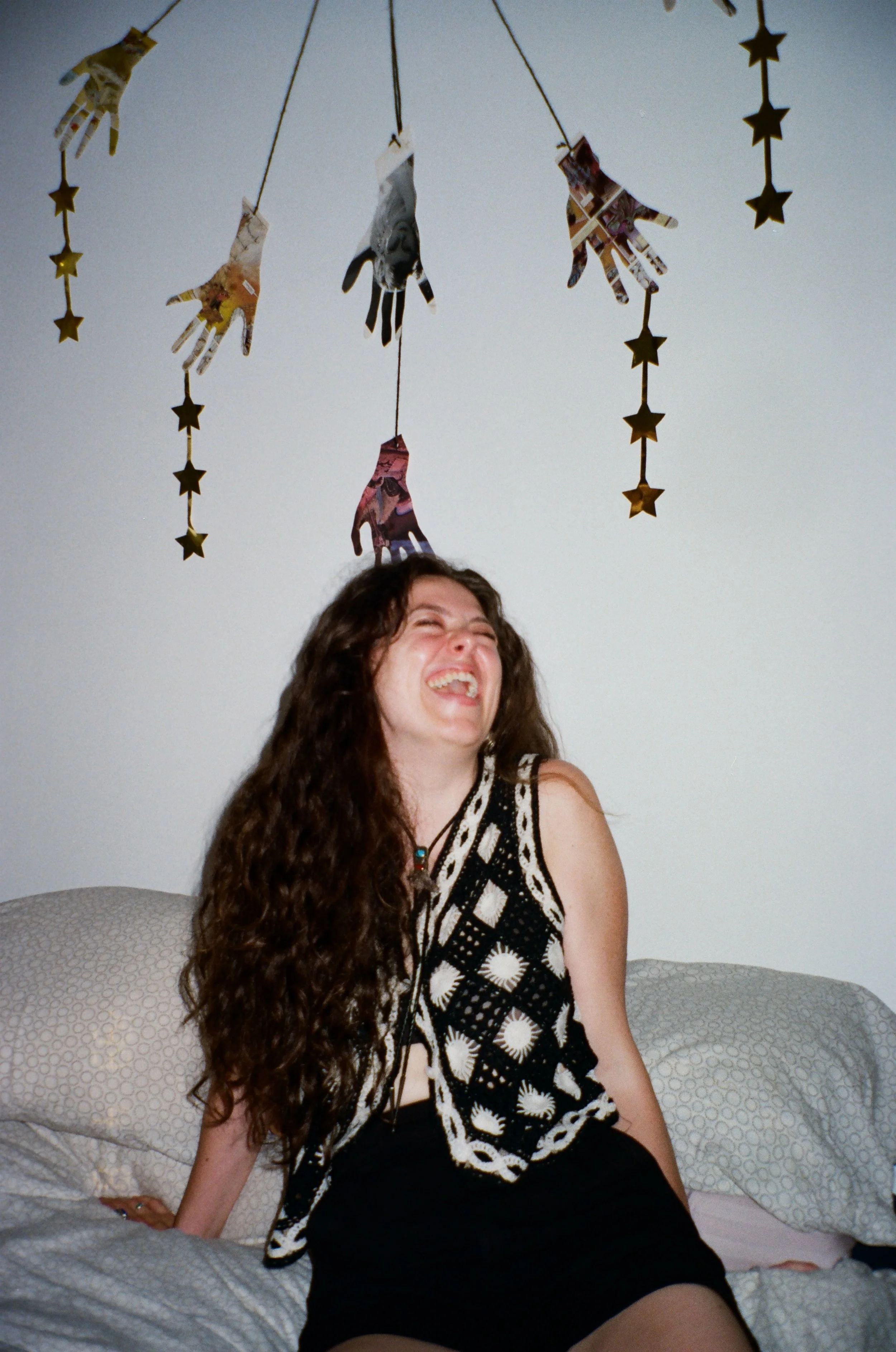 A person with long curly hair laughing while sitting on a bed, with decorative paper hands and star-shaped streamers hanging above her.