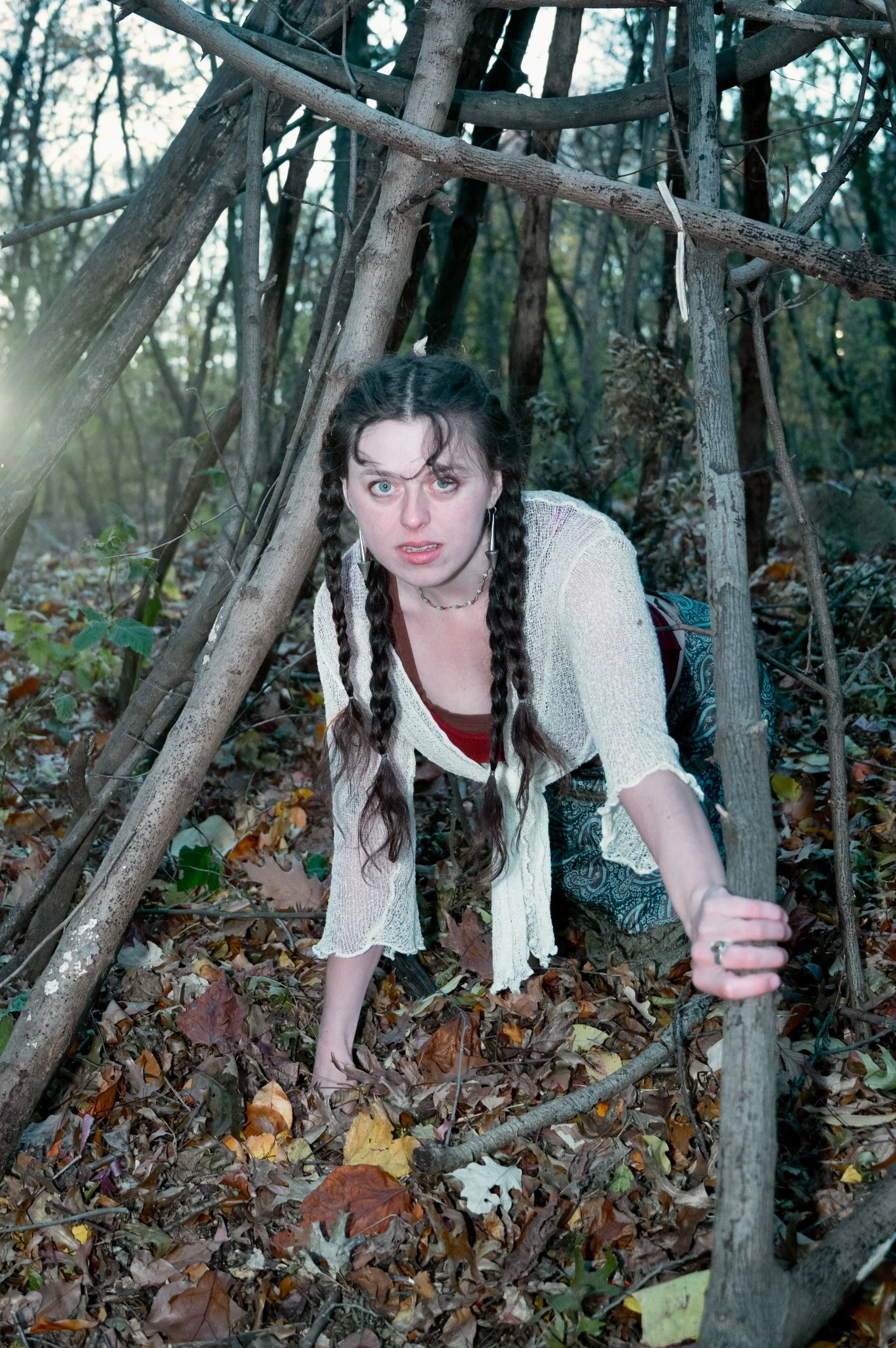 A young person with braided hair crouches on the forest floor, looking up with a startled expression, under a makeshift shelter made of branches and sticks.