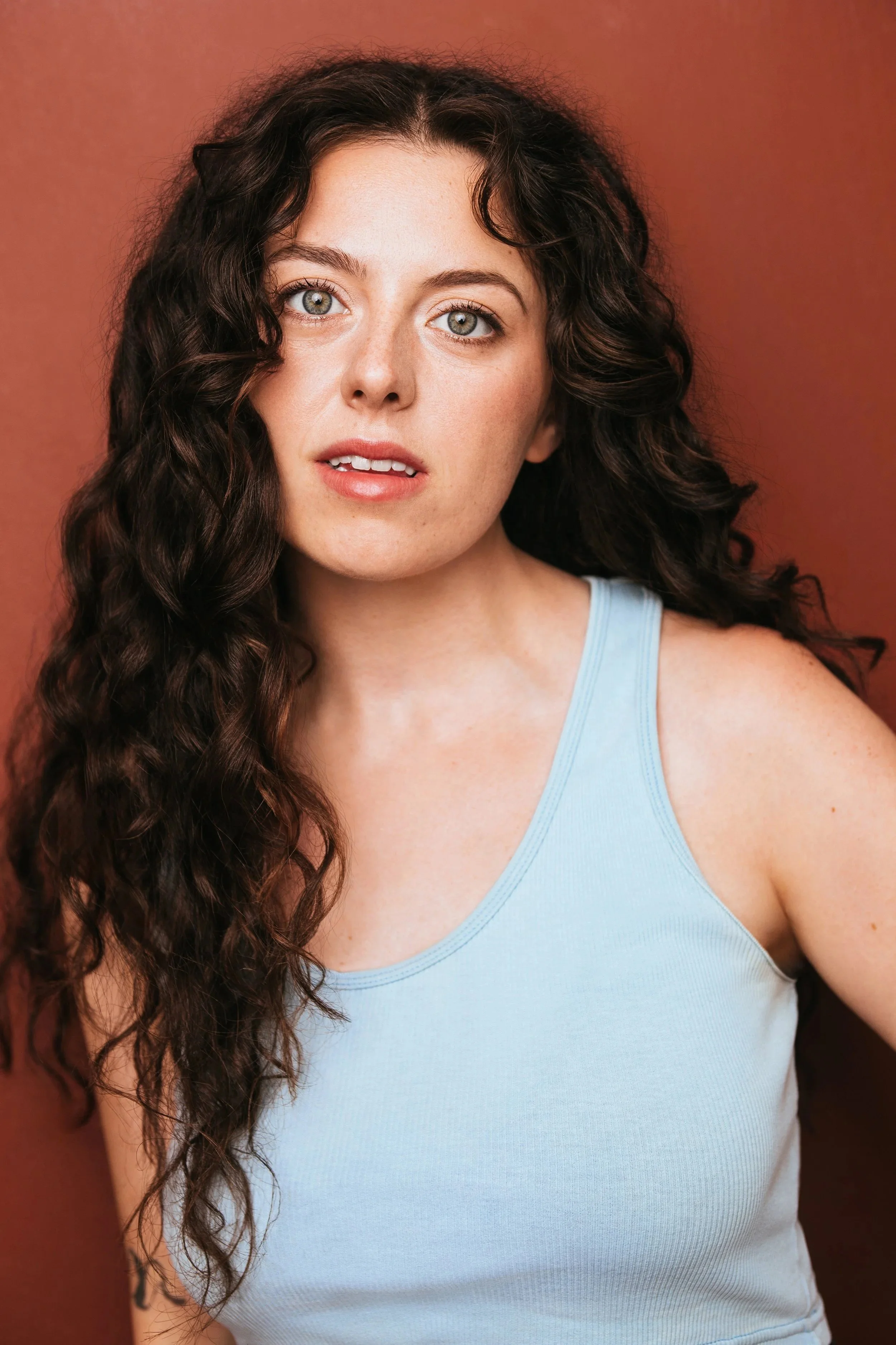 A young person with long, curly dark hair and blue eyes, wearing a light blue tank top, standing against a plain brown background.