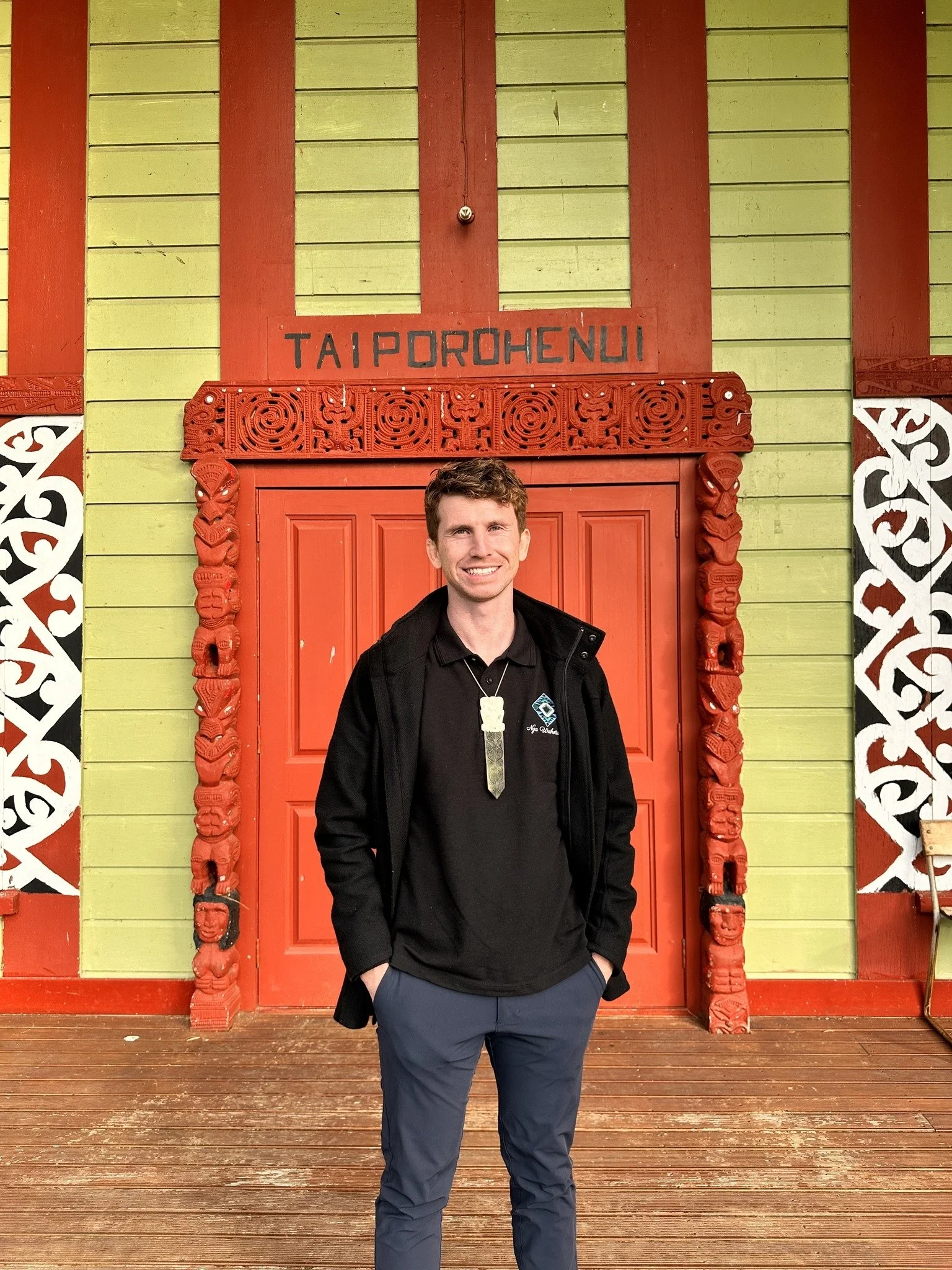 Airana Ngarewa, wearing a black jacket and blue pants, stands on a wooden deck in front of a vibrant green and red wharenui with traditional Māori carvings and the word 'TAIPOROHENUI' on top.