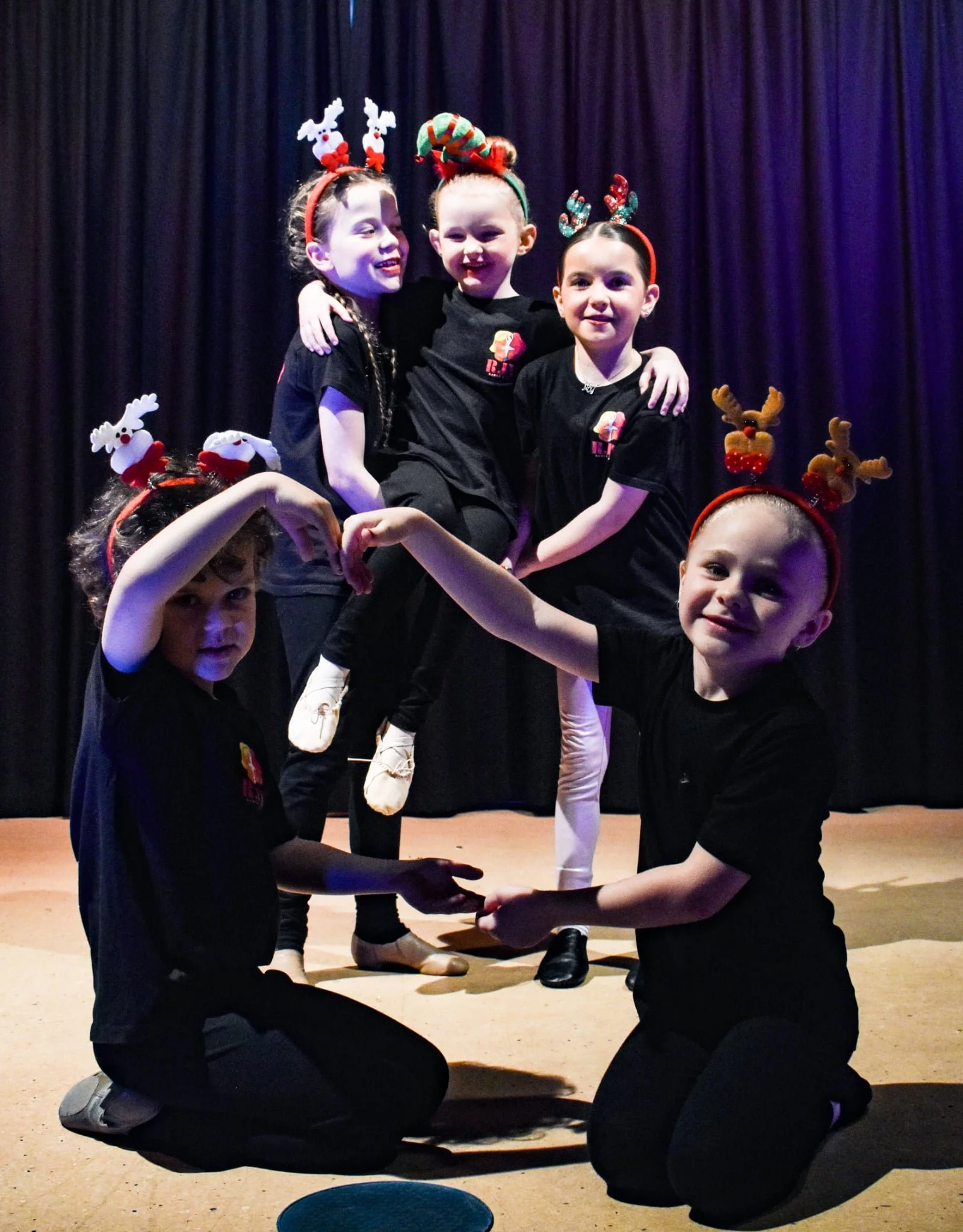 Six children on stage dressed in black, wearing Christmas-themed headbands, with some standing on each other's legs in a formation, posing for a group photo.