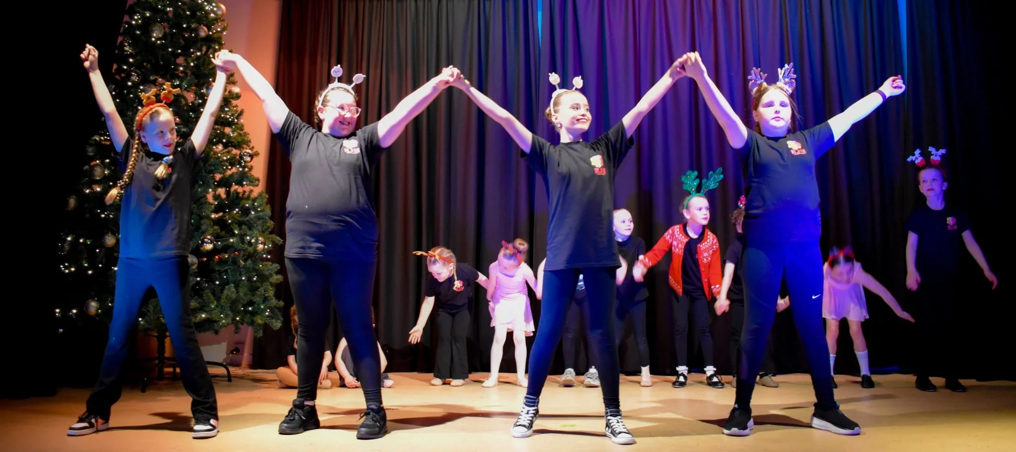 Children and adults dressed in black shirts and Christmas accessories, performing a dance or skit on stage with a decorated Christmas tree on left and a dark curtain backdrop.