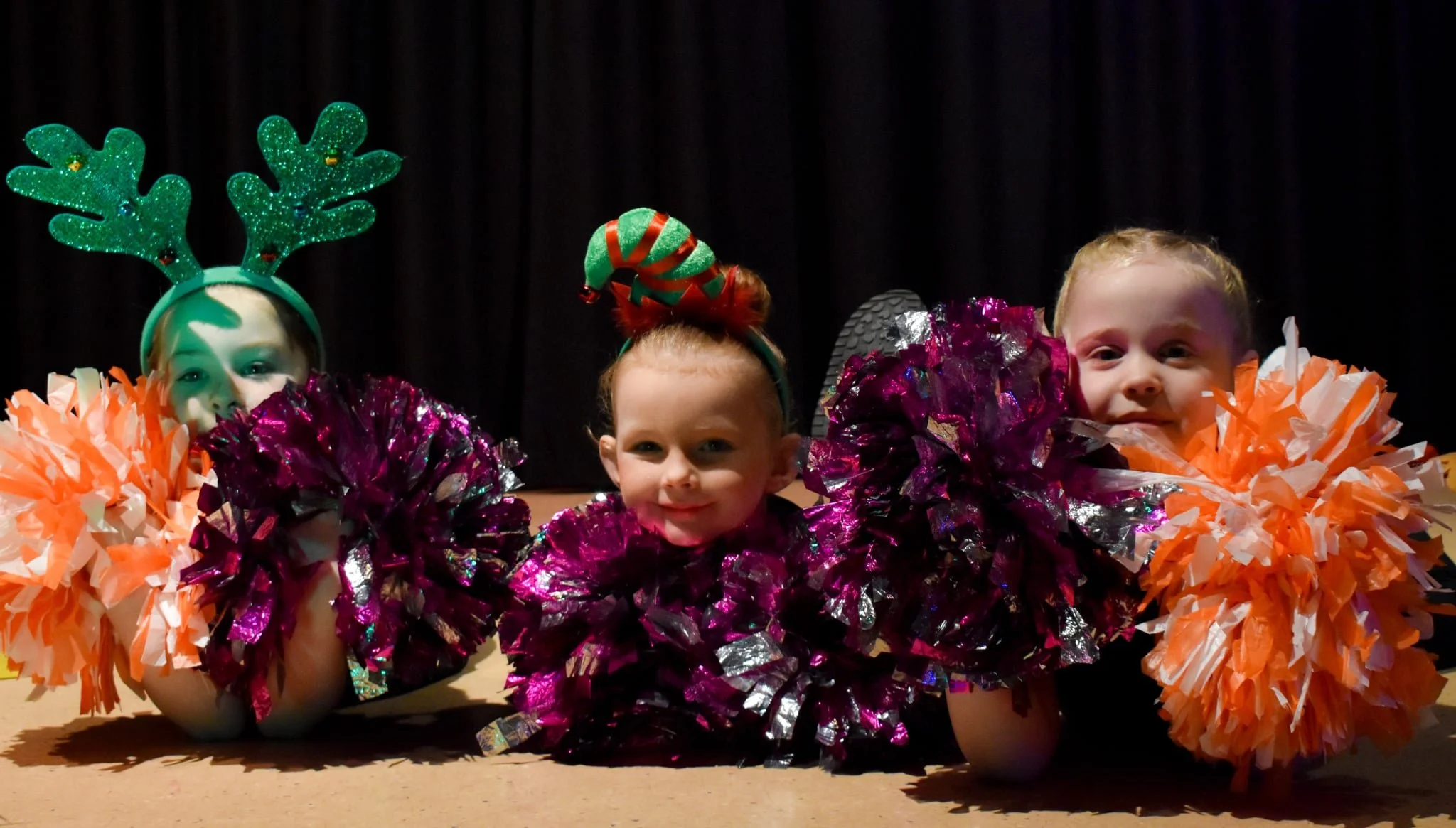 Three young children lying on the floor in festive costumes with colorful pom-poms around their necks, against a dark background.