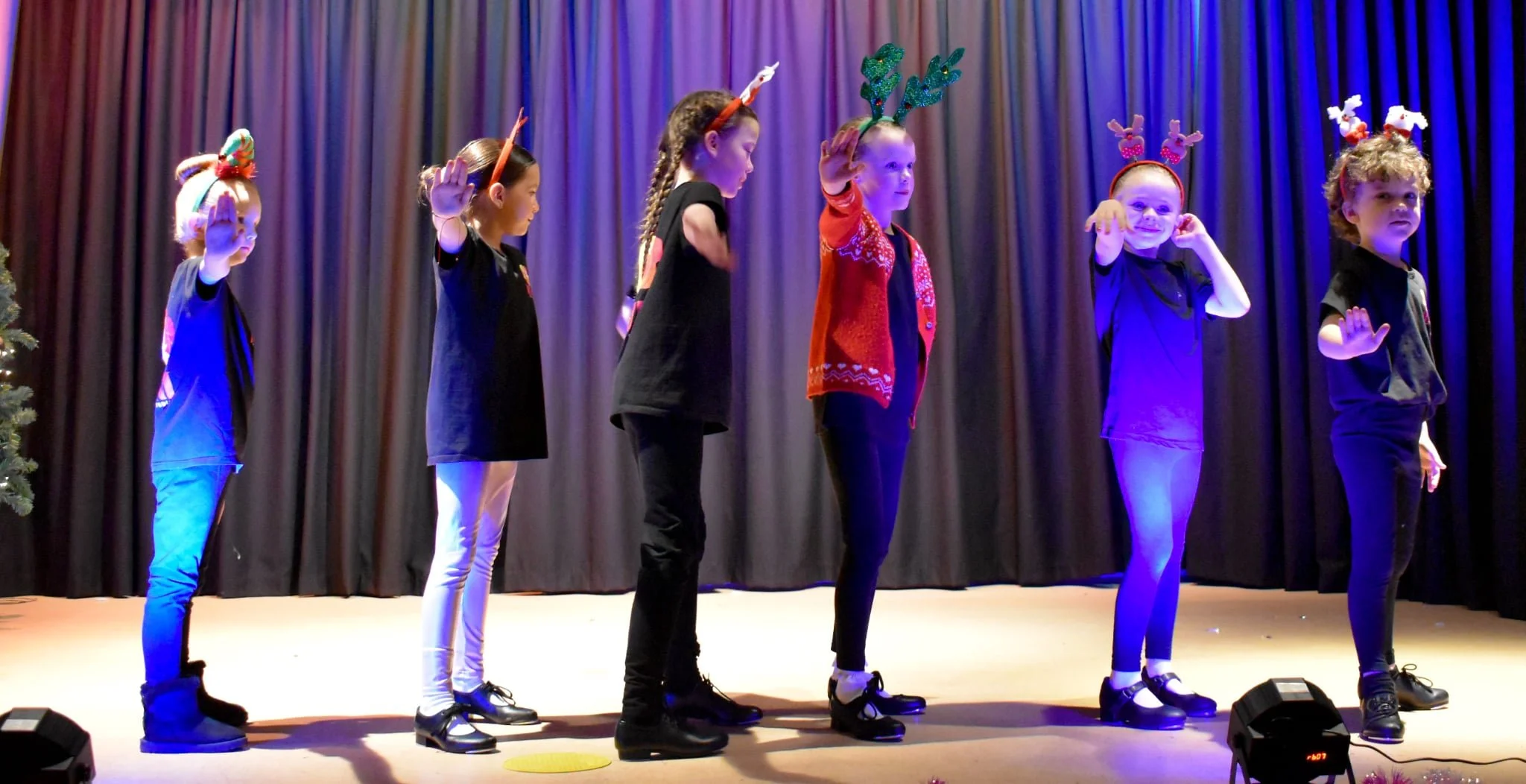 Children dressed in black with reindeer antler headbands and festive accessories perform on a stage with black curtains during a holiday event.