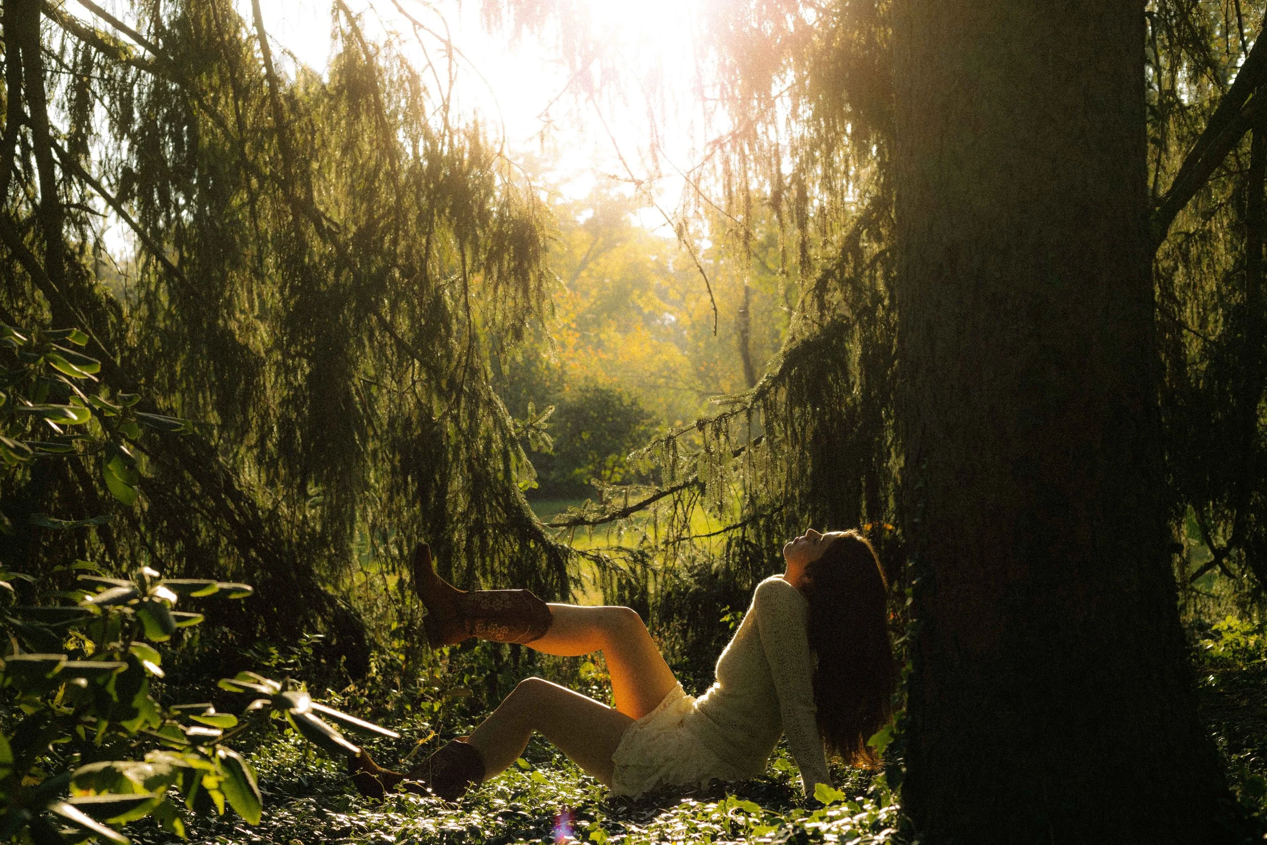 A woman lounging against a tree in a forest at sunset.