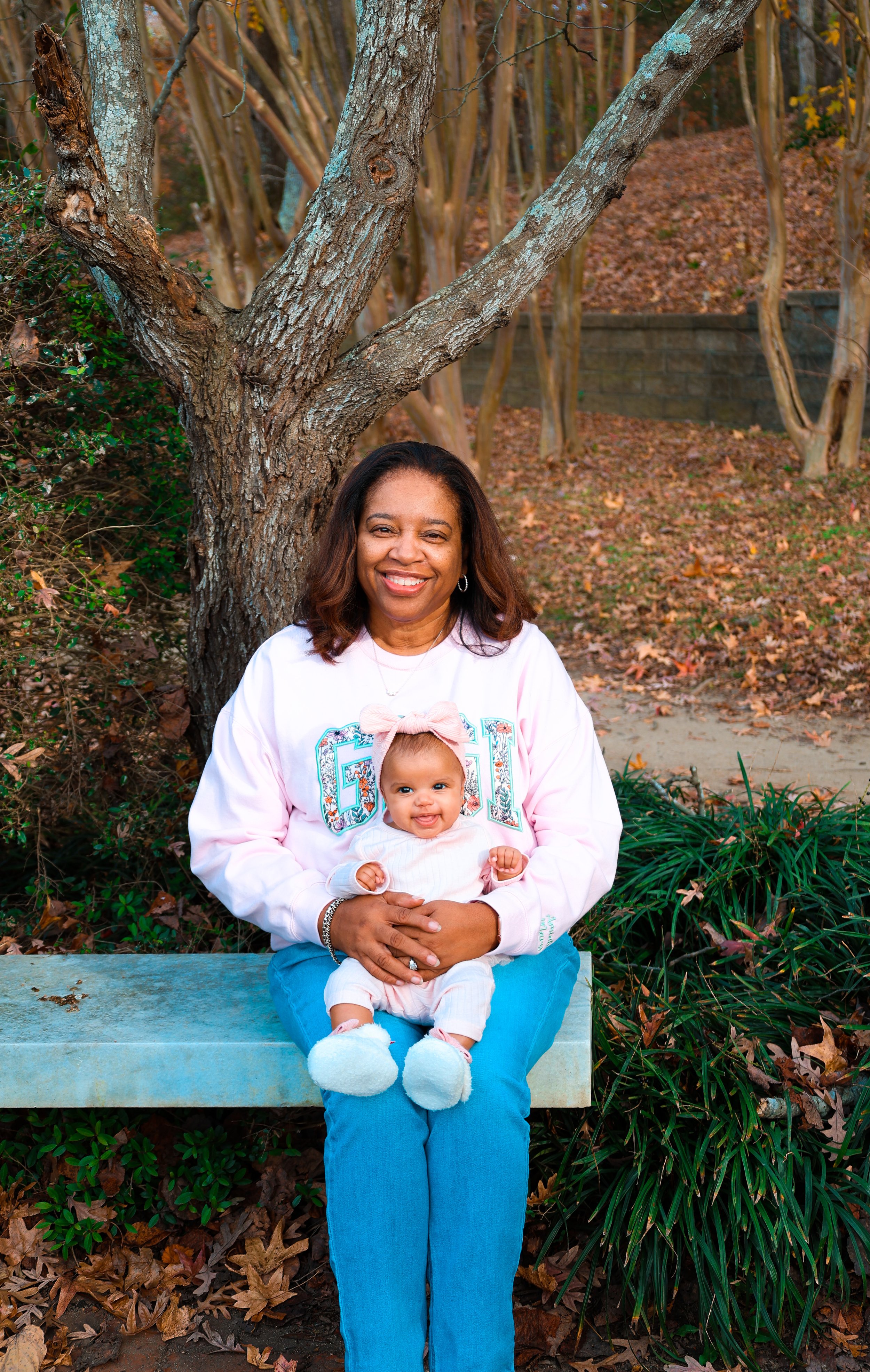 A woman and a baby sitting on a bench outdoors with a tree and fallen leaves in the background. The woman is smiling, wearing a pink sweatshirt and blue jeans, while the baby is dressed in a white outfit with a pink bow headband and fluffy white shoe