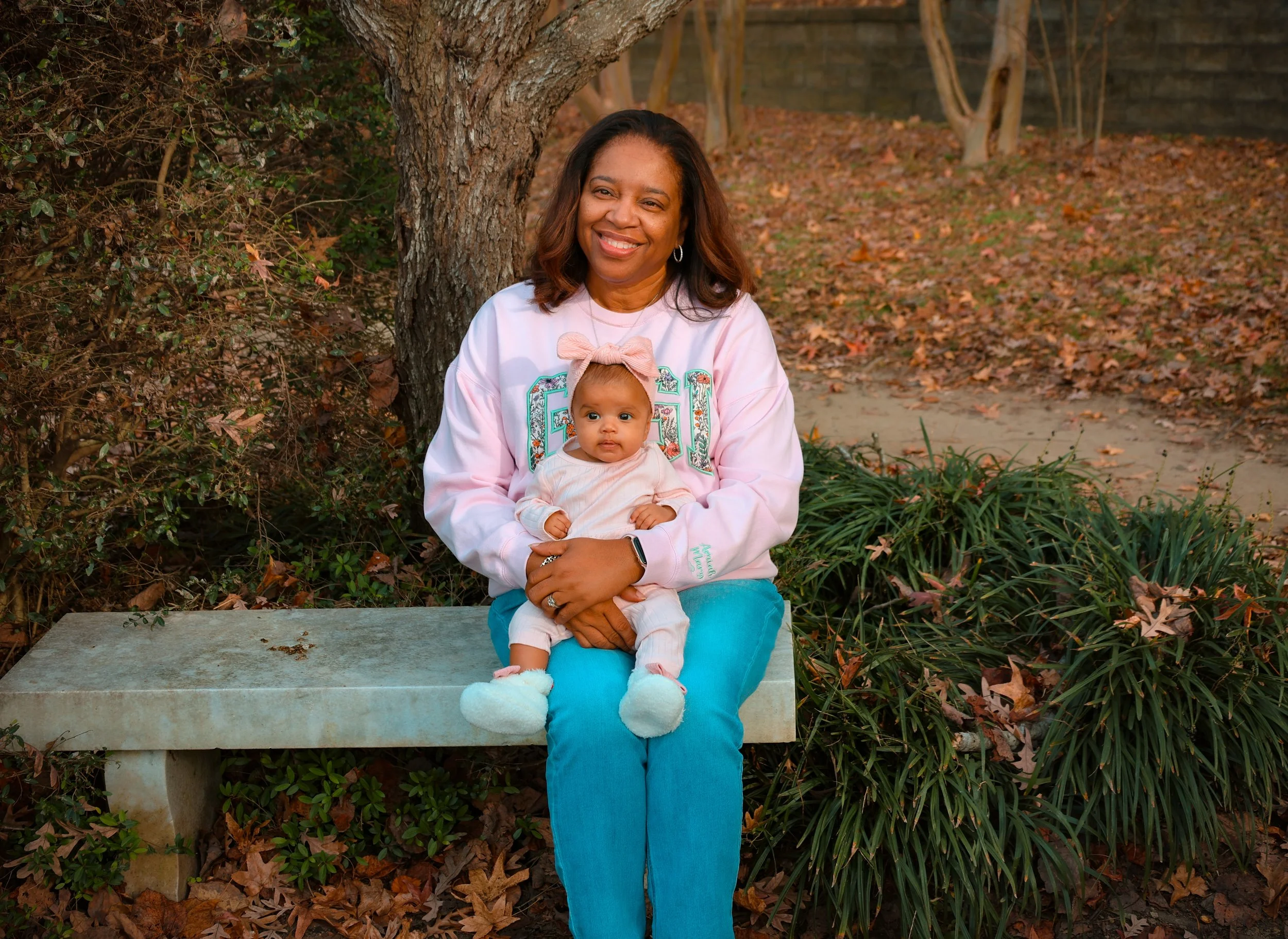 A woman holding a baby girl sits on a concrete bench outdoors surrounded by fall leaves and trees.