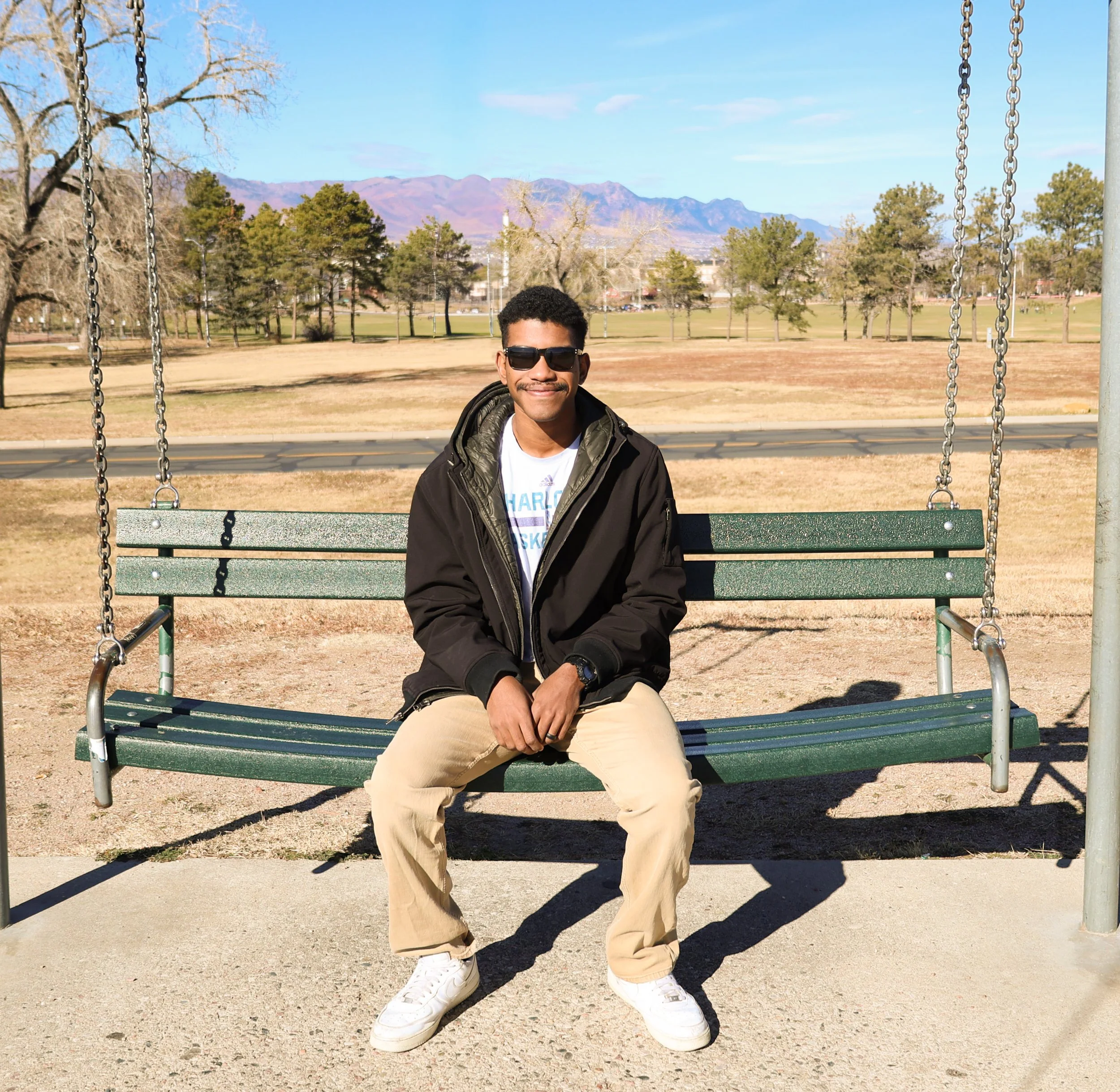 A young man sitting on a park bench swing, wearing sunglasses, a black jacket, a white t-shirt, beige pants, and white sneakers, with a wide smile. The park is open with grass, trees, distant mountains, and a clear blue sky in the background.