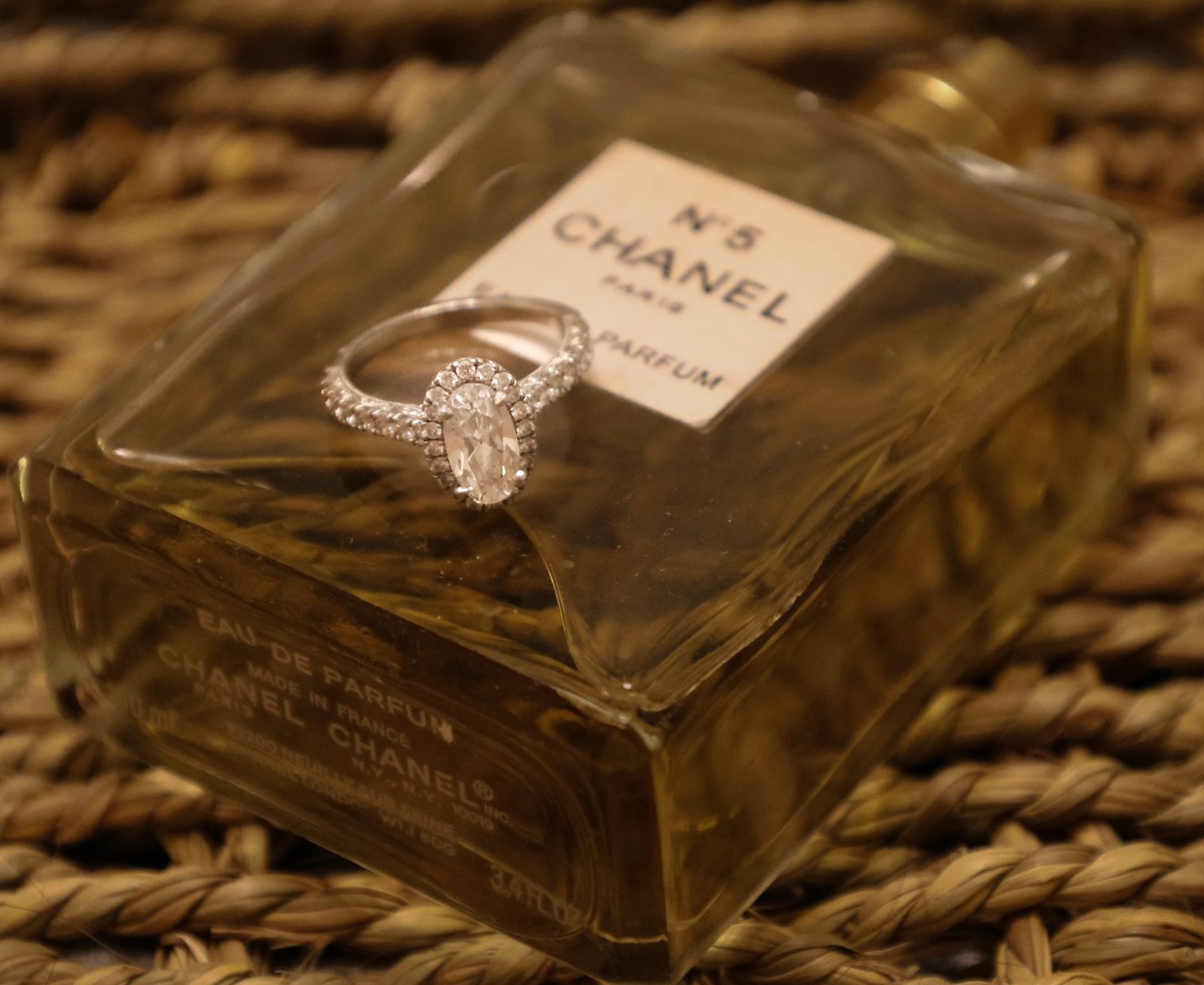 A diamond ring resting on top of a Chanel No. 5 perfume bottle, which is lying on a woven surface.