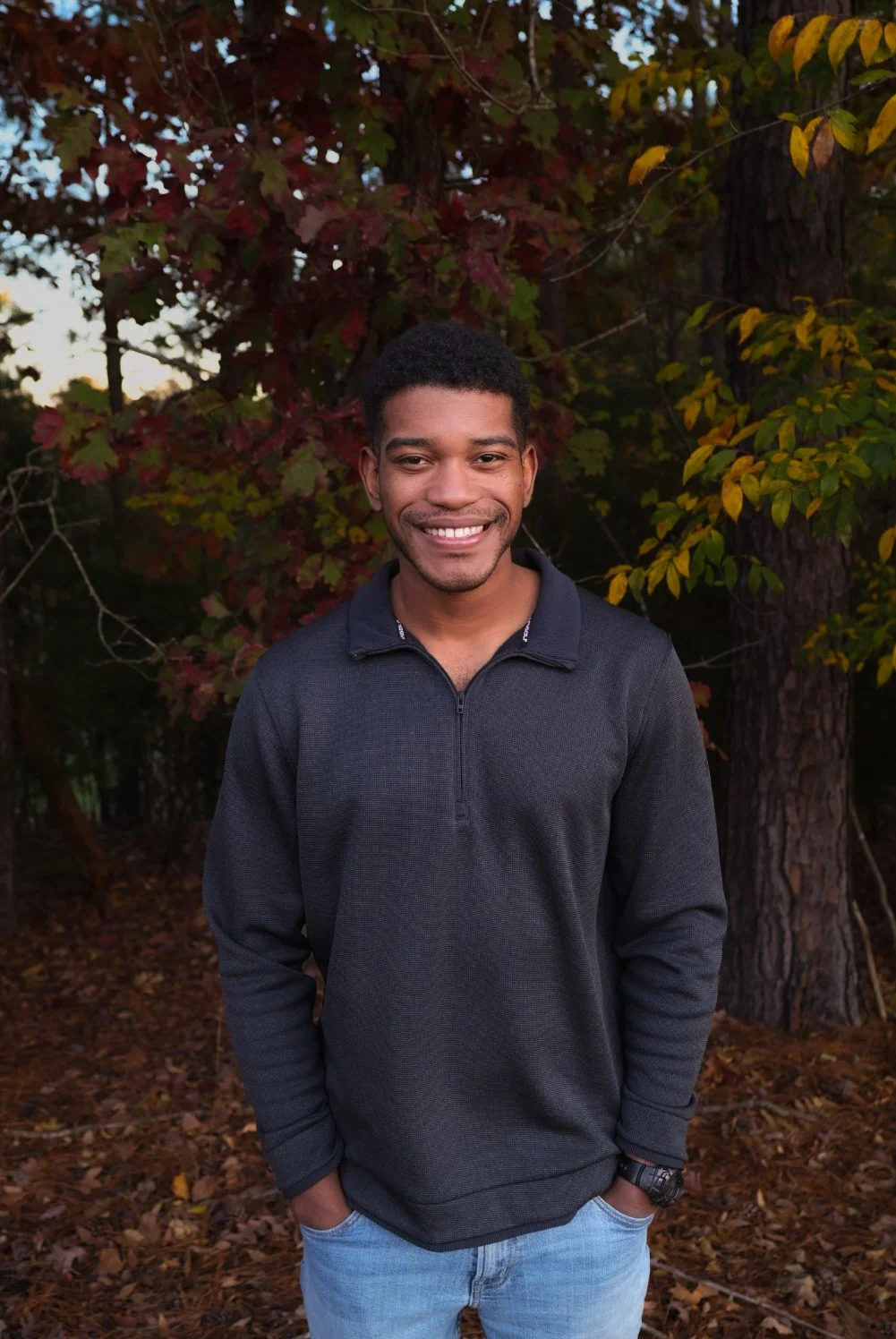 A young man with short black curly hair and dark skin, smiling, standing outdoors in front of trees with autumn-colored leaves. He is wearing a dark gray quarter-zip sweater, light blue jeans, and a black watch on his left wrist.