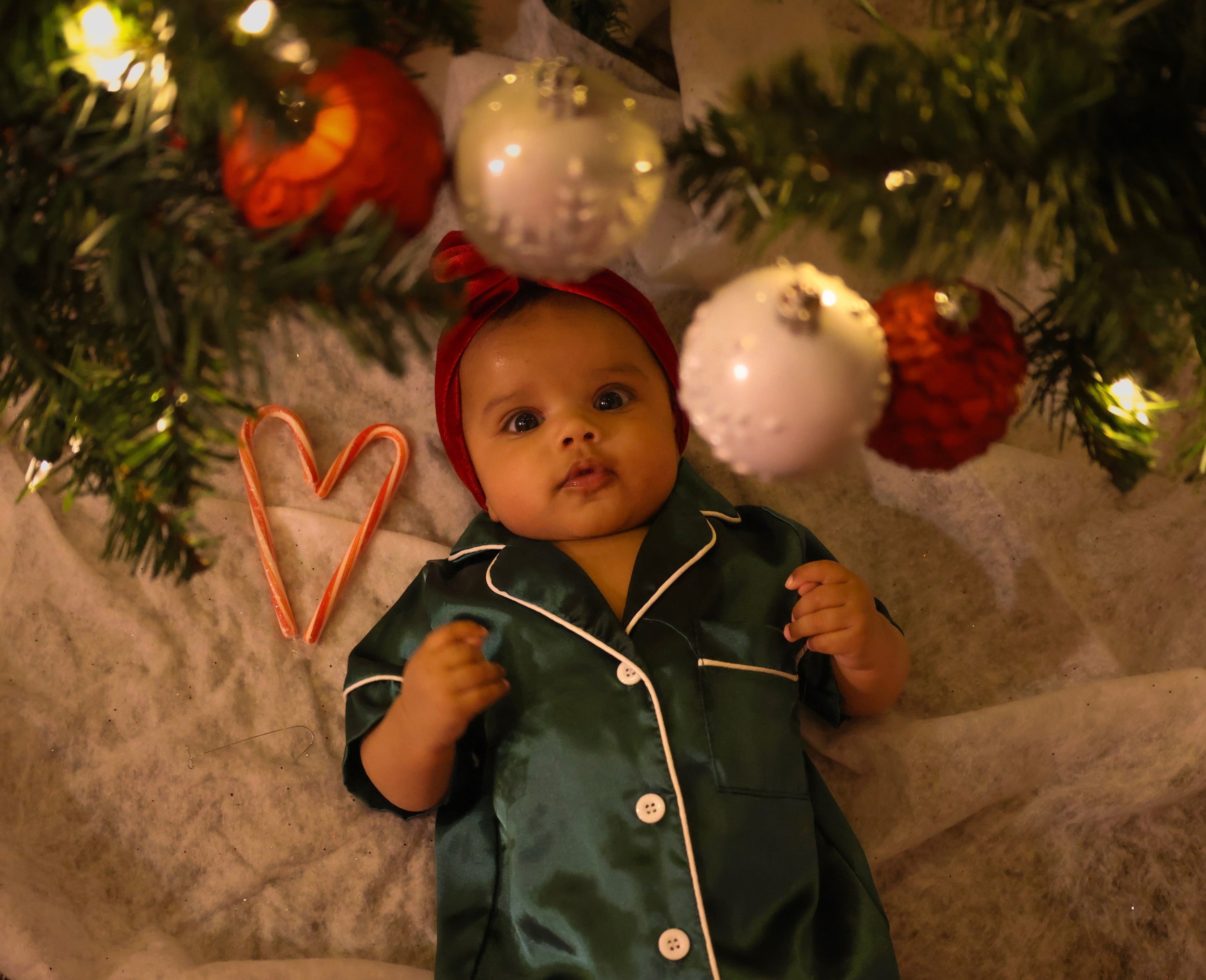 A baby wearing green pajamas and a red headband, lying under a decorated Christmas tree with ornaments, next to a candy cane shaped like a heart.