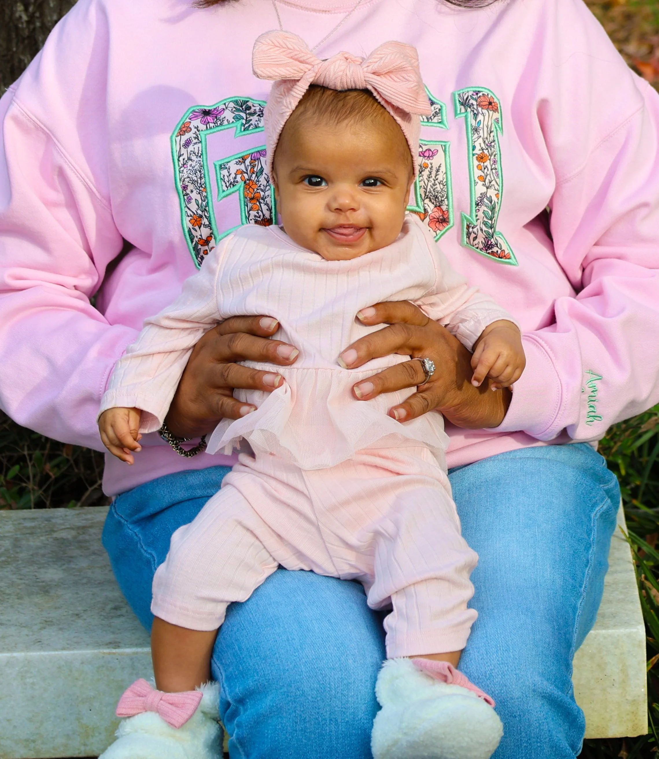 A baby girl sitting on an adult's lap, wearing a pink outfit, a pink bow headband, and pink fluffy shoes, smiling at the camera.