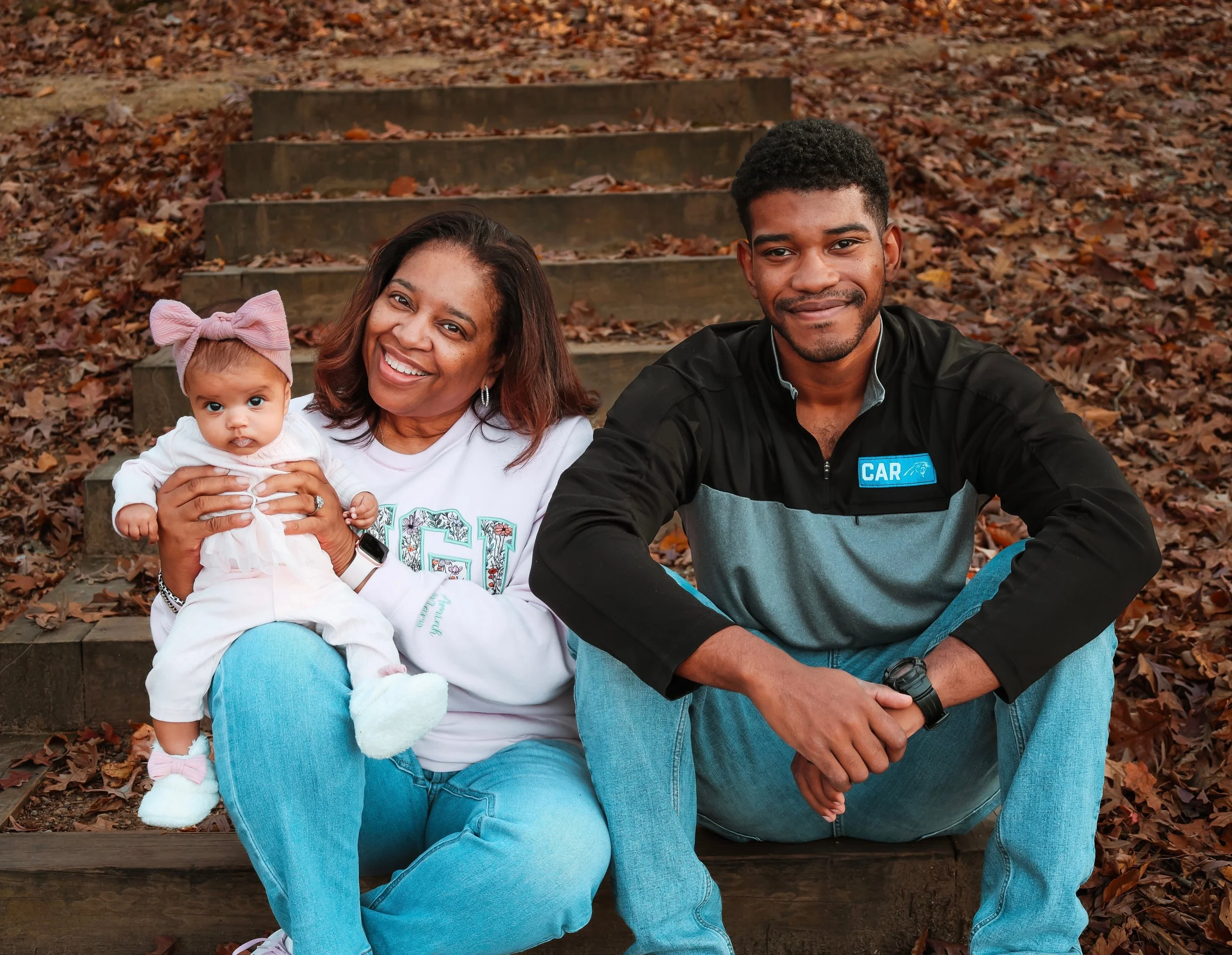 A happy family sitting on outdoor steps covered with autumn leaves. The woman holds a baby girl wearing a pink bow headband, while the man sits next to them, smiling at the camera.