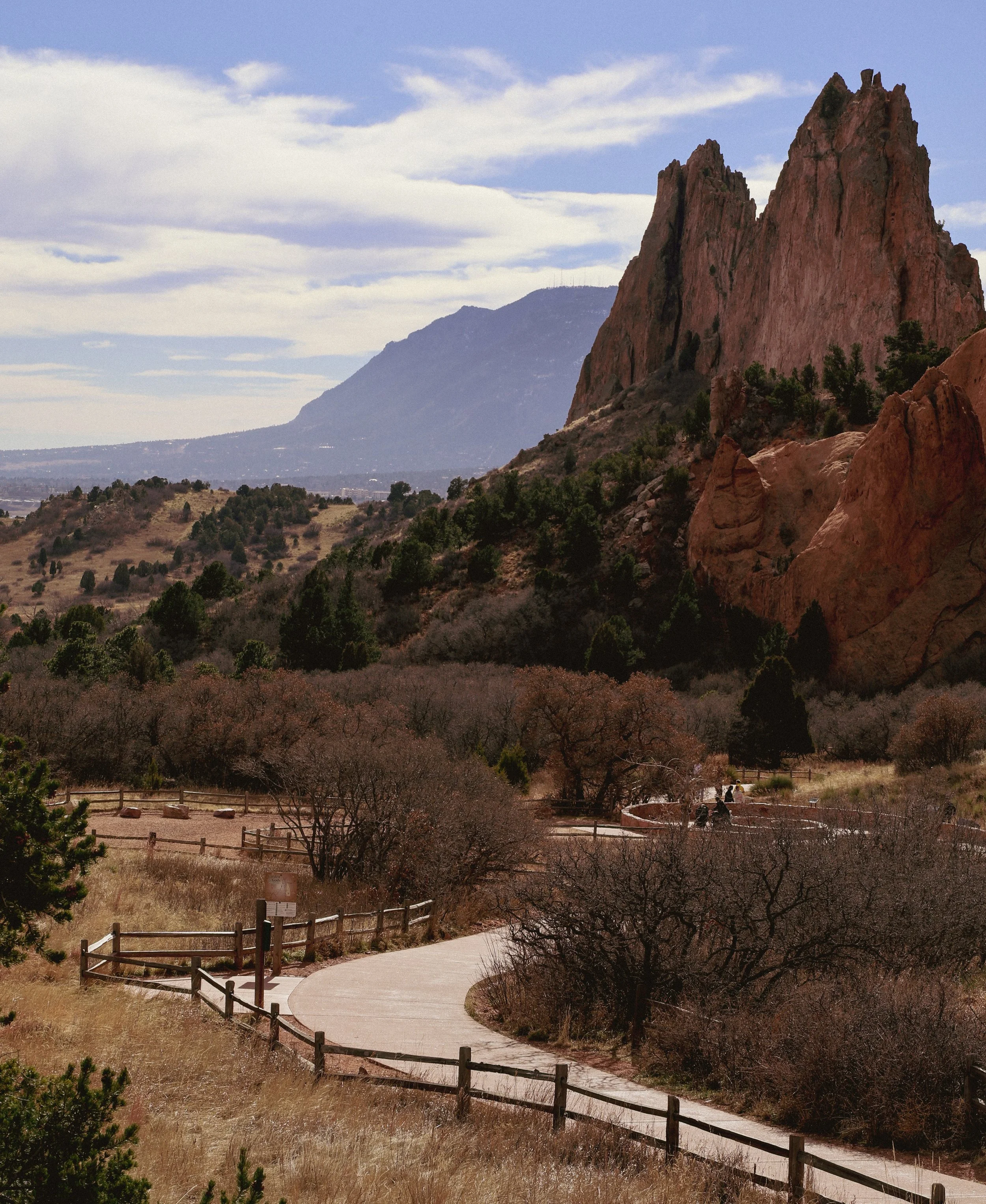 A winding pathway through a dry, hilly landscape with sparse trees, leading towards large red rock formations and distant mountains under a partly cloudy sky.