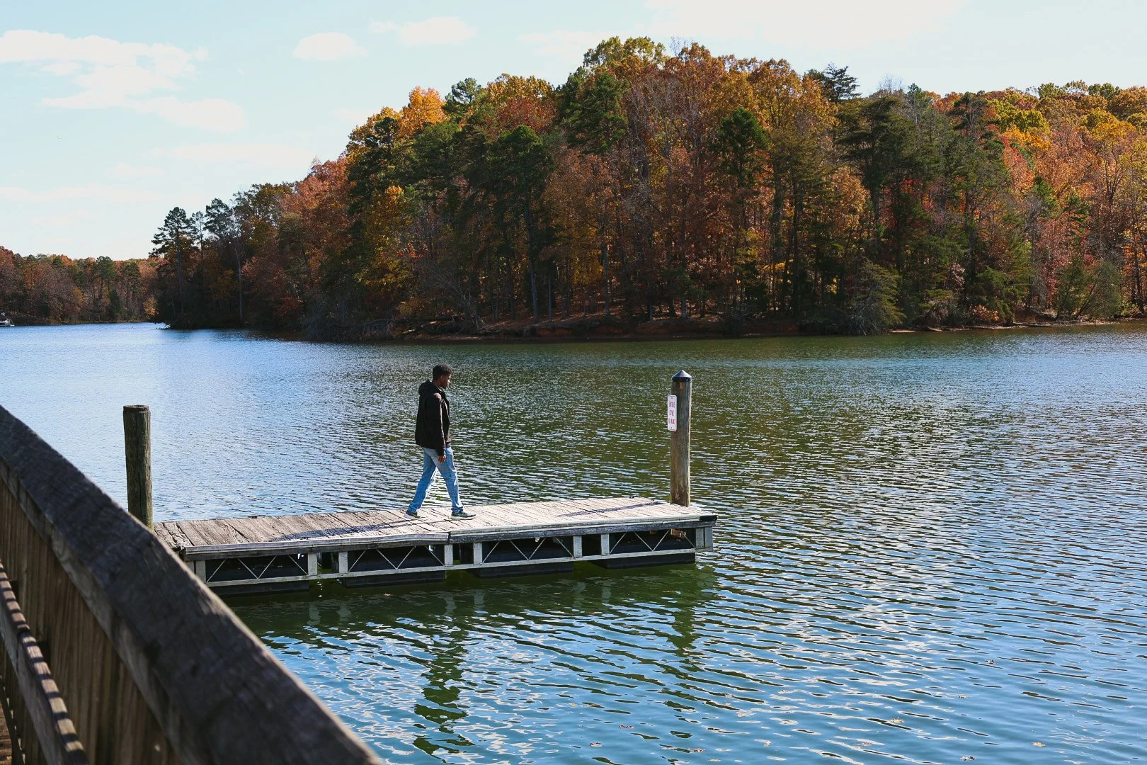 A person walking on a wooden dock by a lake with trees displaying fall foliage in the background.