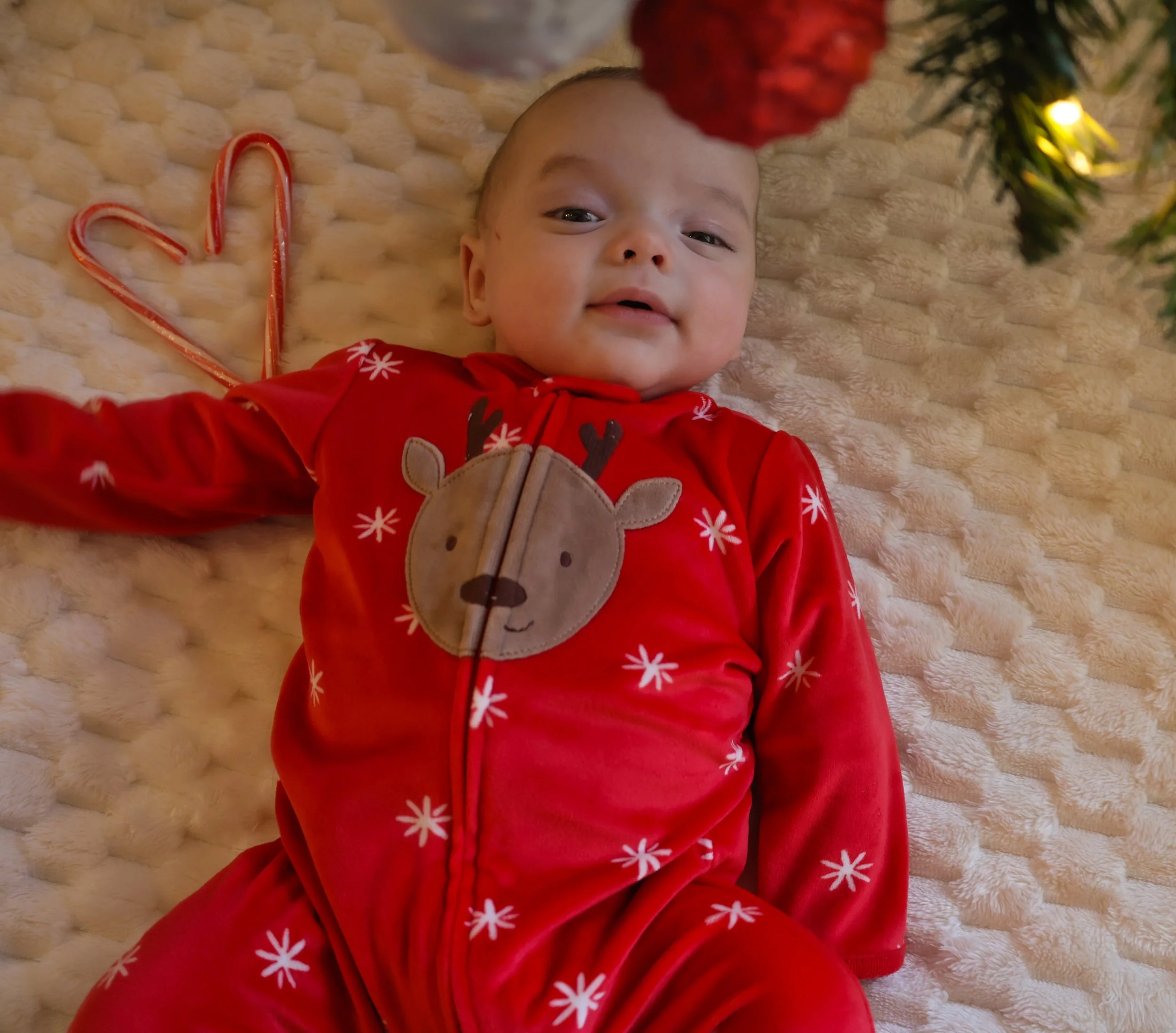 A baby lying on a soft, cream-colored textured blanket, wearing a red Christmas-themed onesie with a reindeer face on the front, surrounded by holiday decorations including a heart-shaped candy cane and Christmas tree with lights.