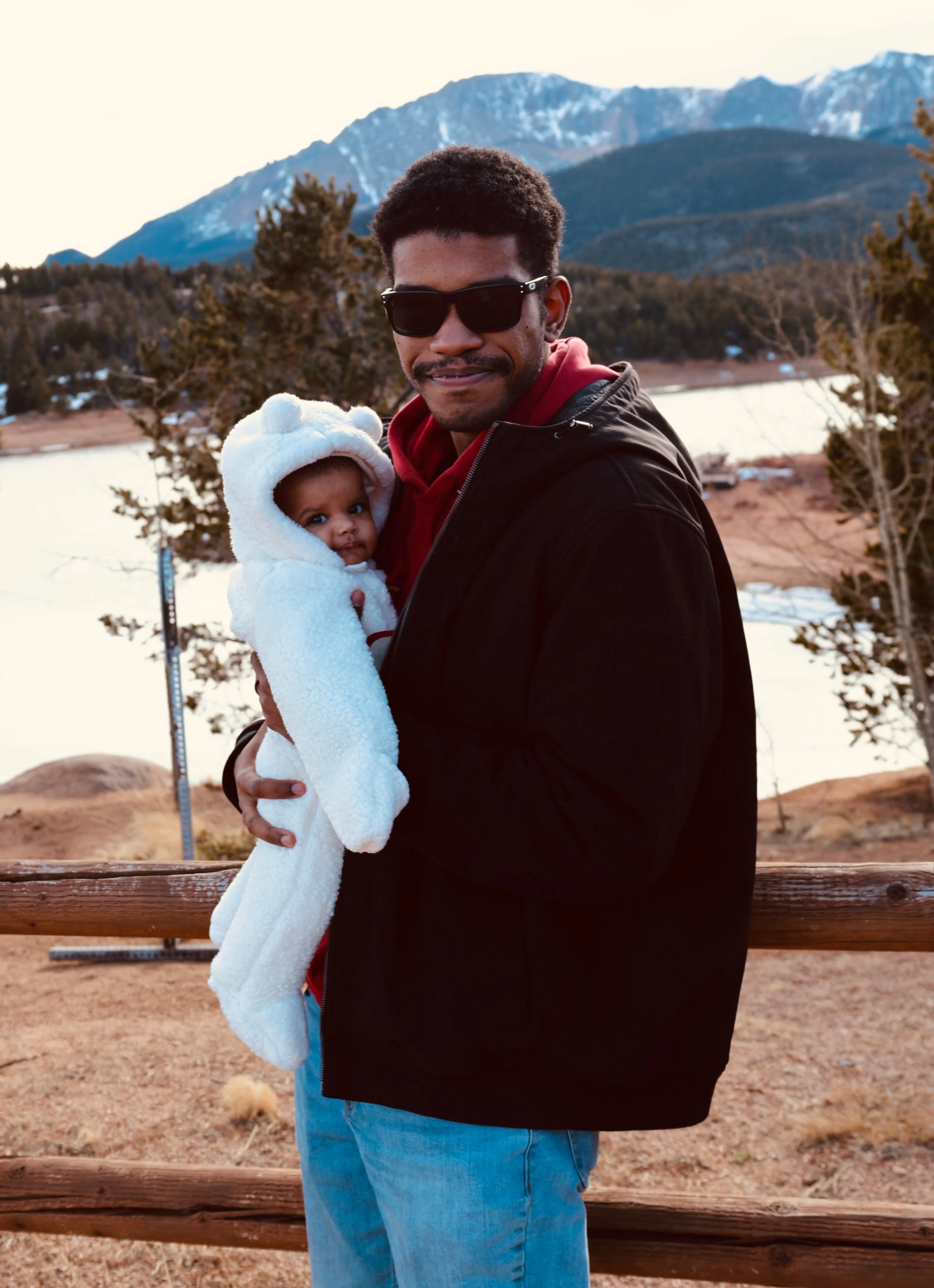 A man wearing sunglasses and casual clothing holding a baby dressed in a cozy white bear costume outdoors near a lake and mountains.