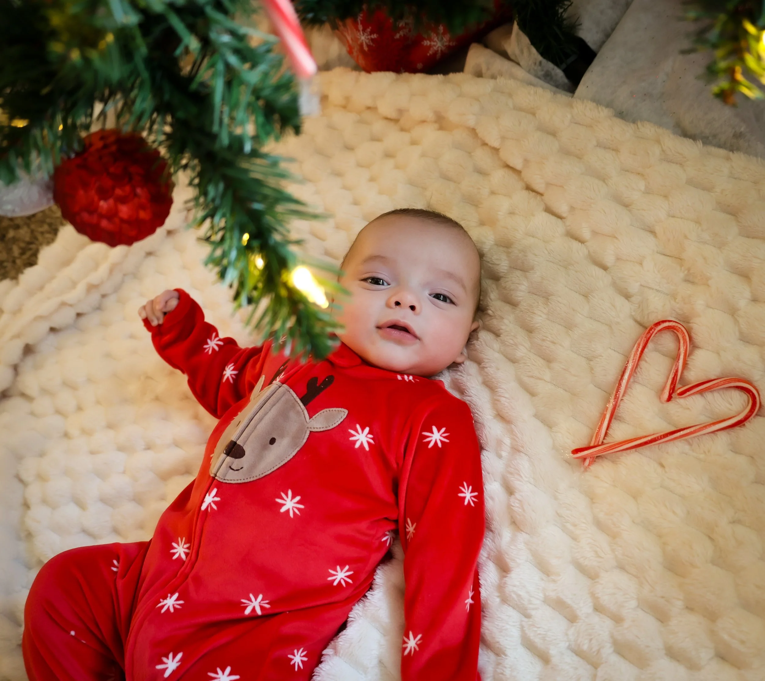 A baby in red Christmas pajamas with reindeer and snowflake patterns lying on a plush cream-colored blanket, under a decorated Christmas tree with ornaments and lights, with two candy canes forming a heart shape nearby.