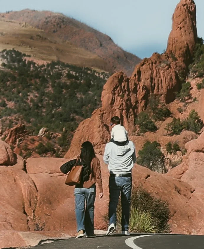 A family of three hiking in a red rock canyon. The woman is dressed in a brown jacket and jeans, carrying a backpack. The man is wearing a white hoodie and jeans, with a child on his shoulders. The landscape features red rock formations and sparse gr