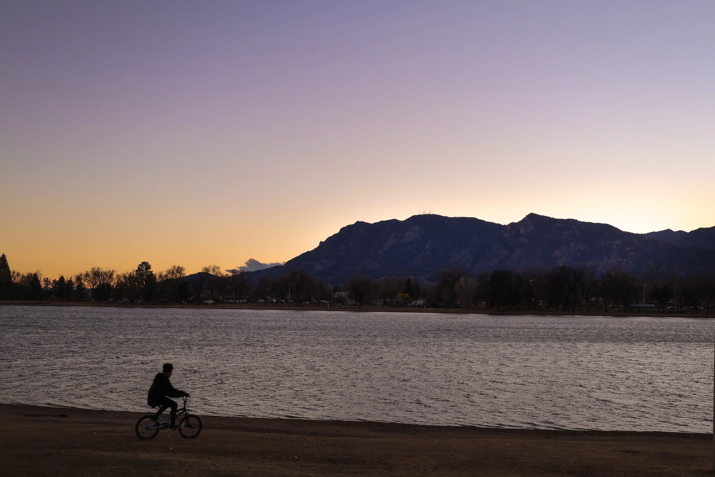 A person riding a bicycle along the shoreline of a lake during sunset, with mountains in the background and a sky transitioning from orange to purple.
