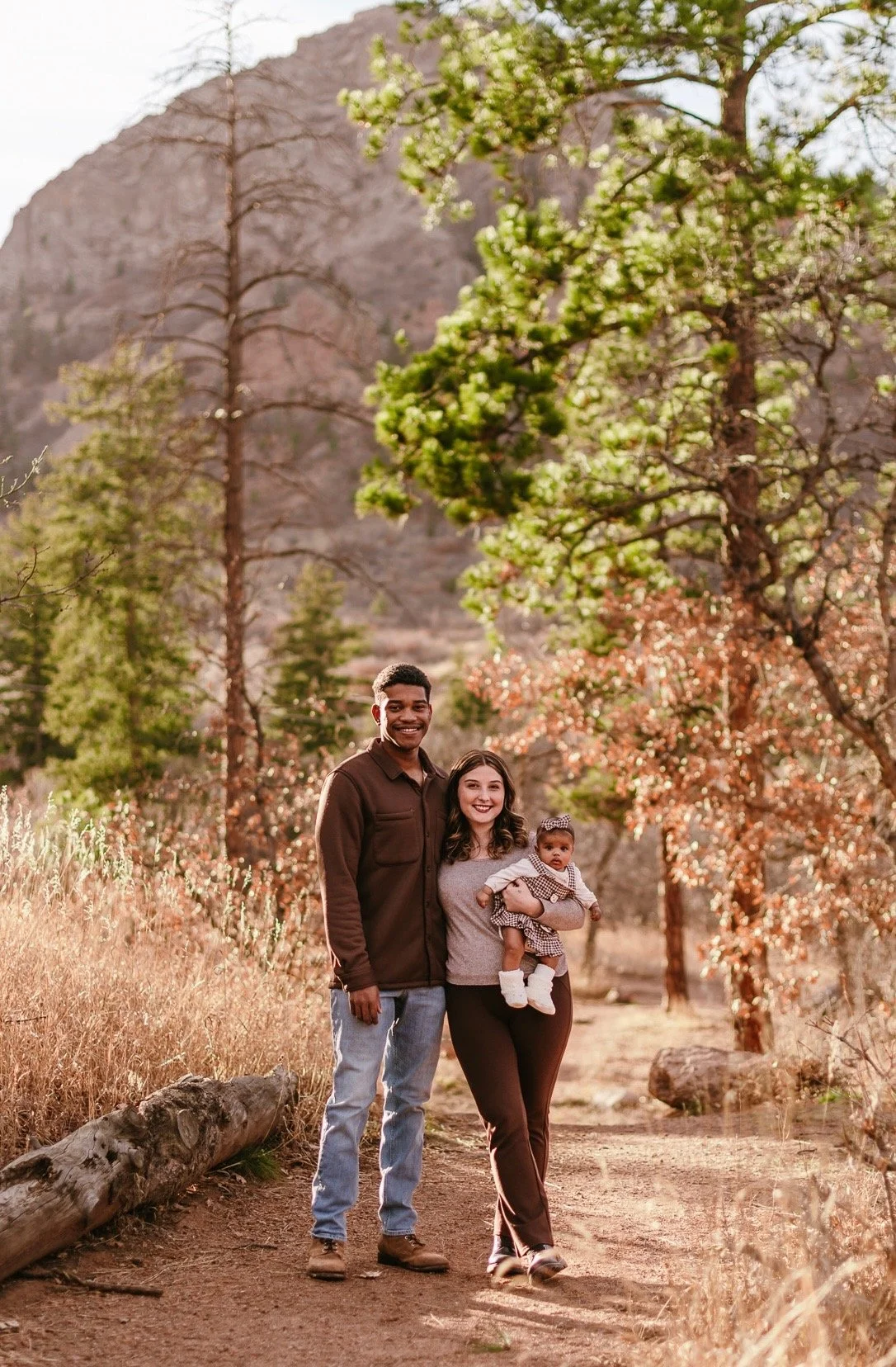 A family of three smiling and walking on a dirt trail in a mountainous area with trees and dry grass.