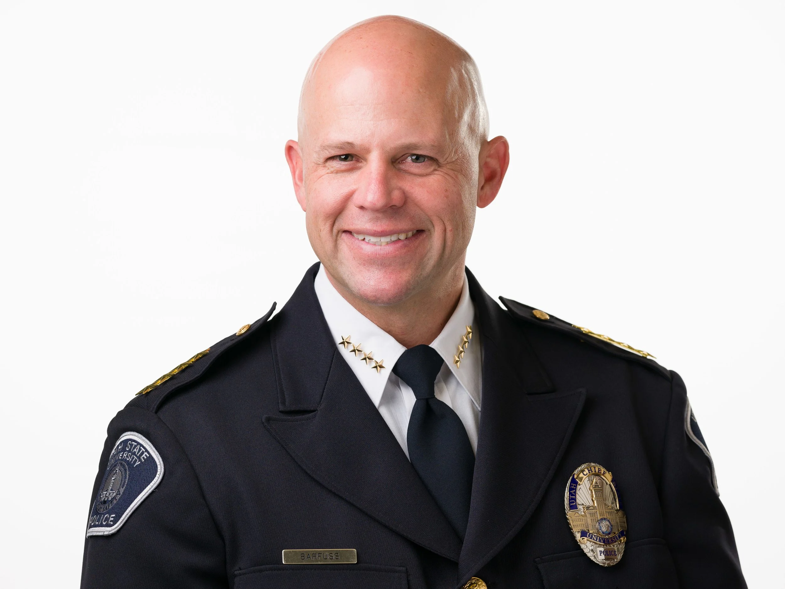 Portrait of Chief Blair Barfuss in a class A police uniform jacket with gold stars on collar and shoulders, white dress shirt, blue tie, and dark blue police dress jacket.