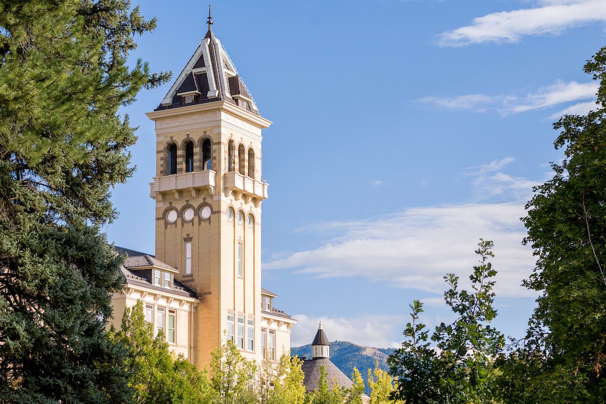 A historic courthouse tower with a pointed roof, beige brick walls, and arched windows, surrounded by green trees under a partly cloudy blue sky.