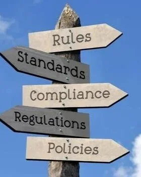 Signpost with arrows pointing in different directions labeled Rules, Standards, Compliance, Regulations, Policies, set against a blue sky.