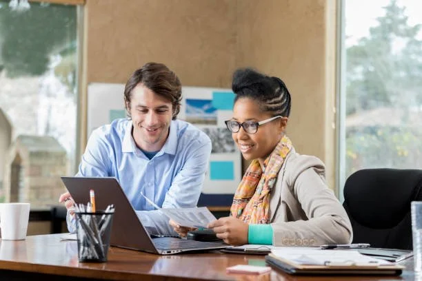 Two people, a man and a woman, sitting at a desk with a laptop, working together in an office setting.