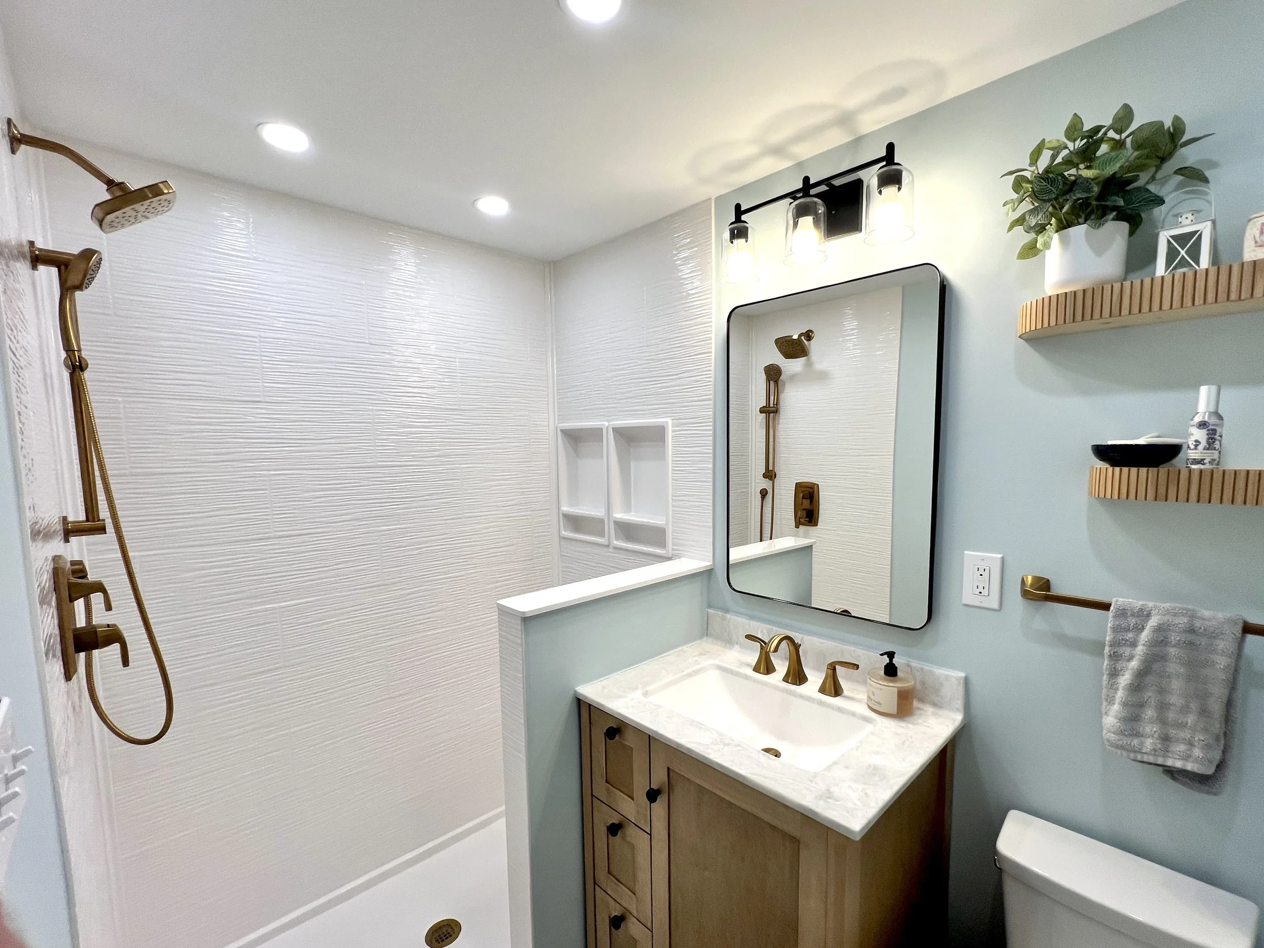 Bathroom with shower area featuring white textured wall tiles, a wooden vanity with a marble top, a large mirror, and gold fixtures. Shelves on the wall hold decorative items and toiletries, with a towel hanging on a gold bar.