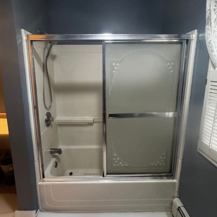 A bathtub with a sliding glass door and a showerhead inside, set in a bathroom with gray walls and a window with white shutters.