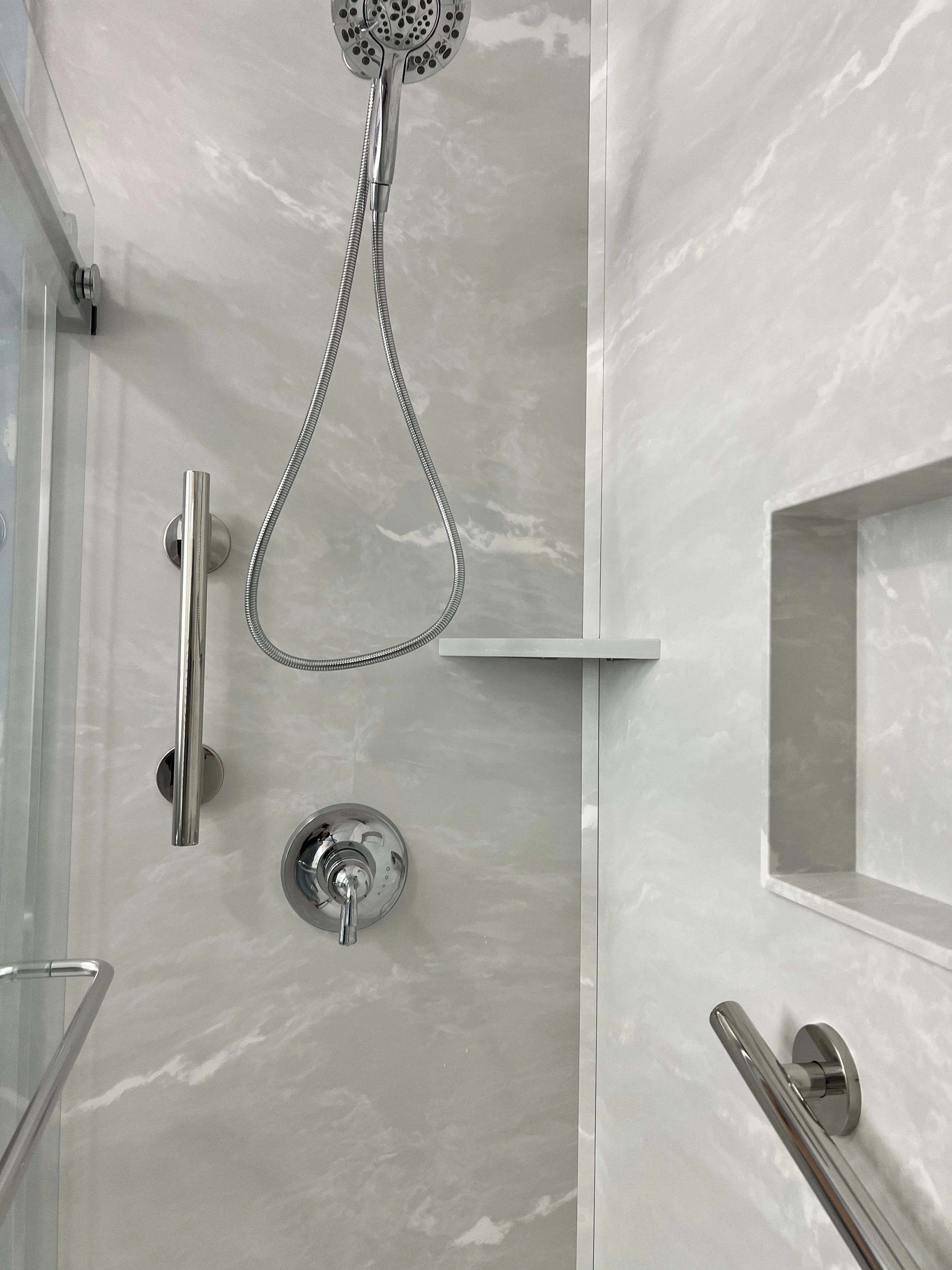 Close-up view of a shower with a handheld showerhead, a control knob, stainless steel grab bar, a built-in shelf, and a glass door.