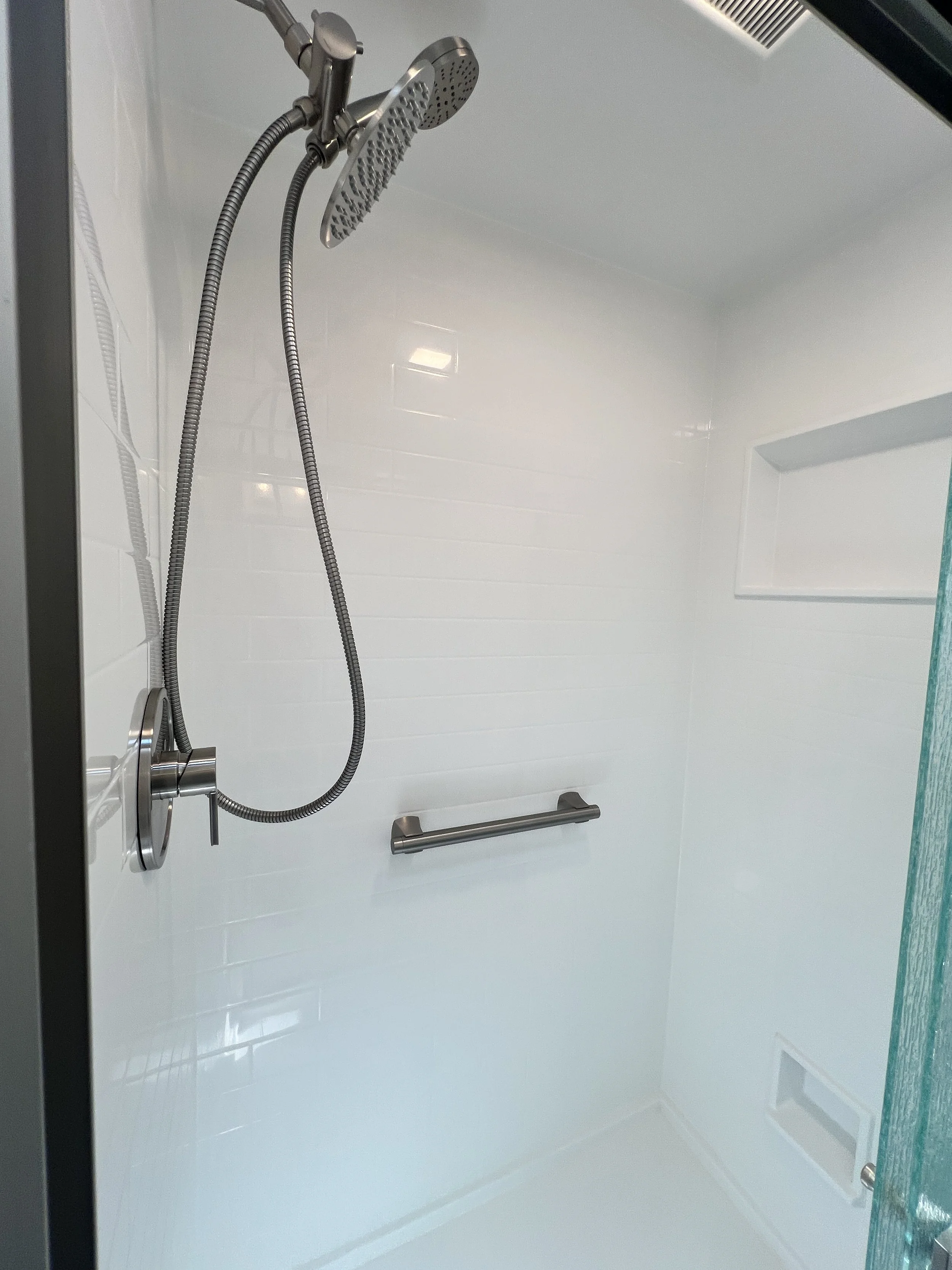 Empty shower with white tiled walls, a wall-mounted silver grab bar, a handheld showerhead attached to a hose, and a built-in soap or shampoo niche.