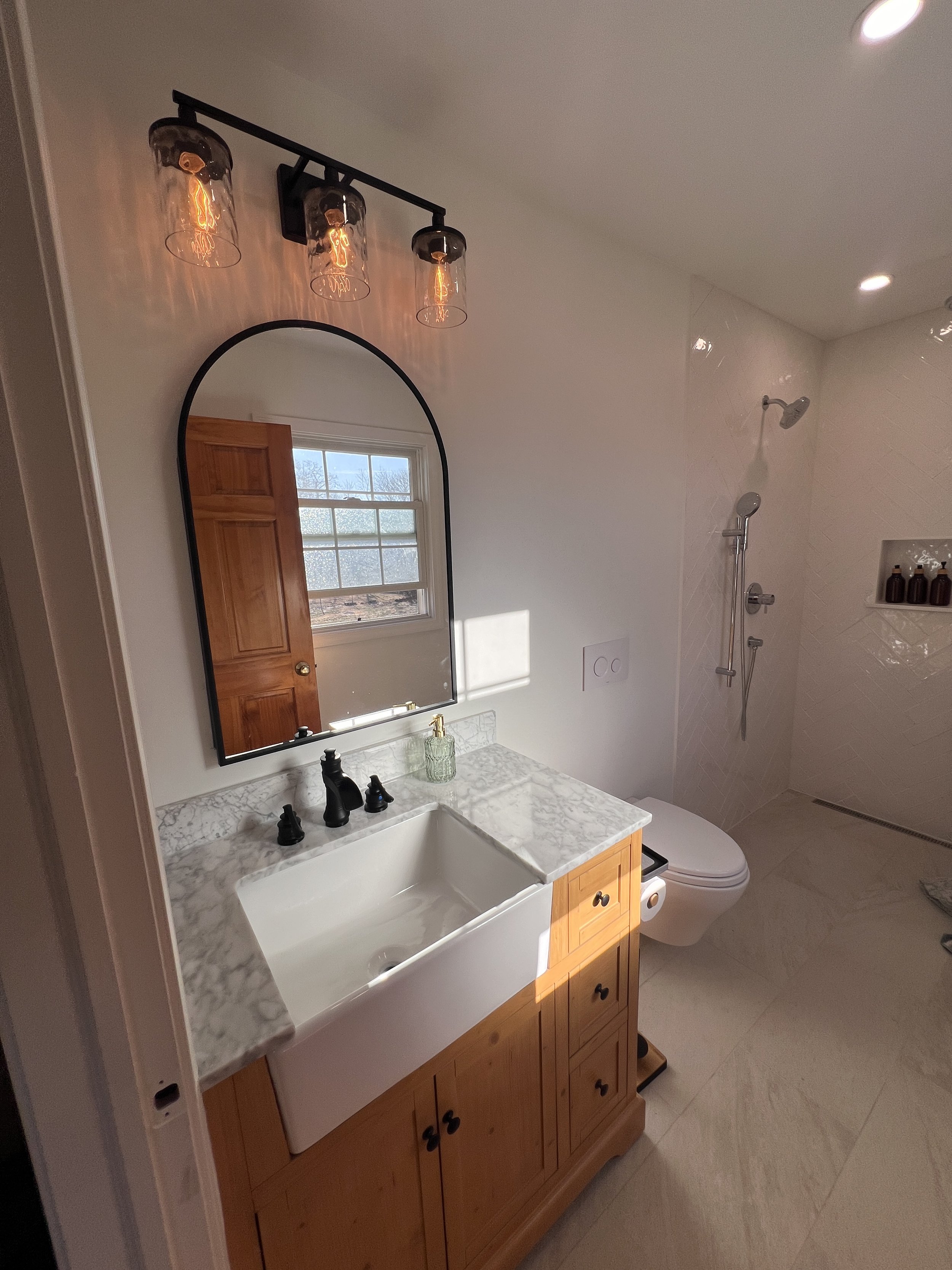 A bathroom with a wooden vanity with a marble countertop, a white apron sink, a black faucet, a mirror, and a wall-mounted light fixture with three exposed bulbs. There is a window with a wooden door reflected in the mirror. The bathroom has a shower area with a wall-mounted showerhead, a niche with bottles, and a beige tiled floor.
