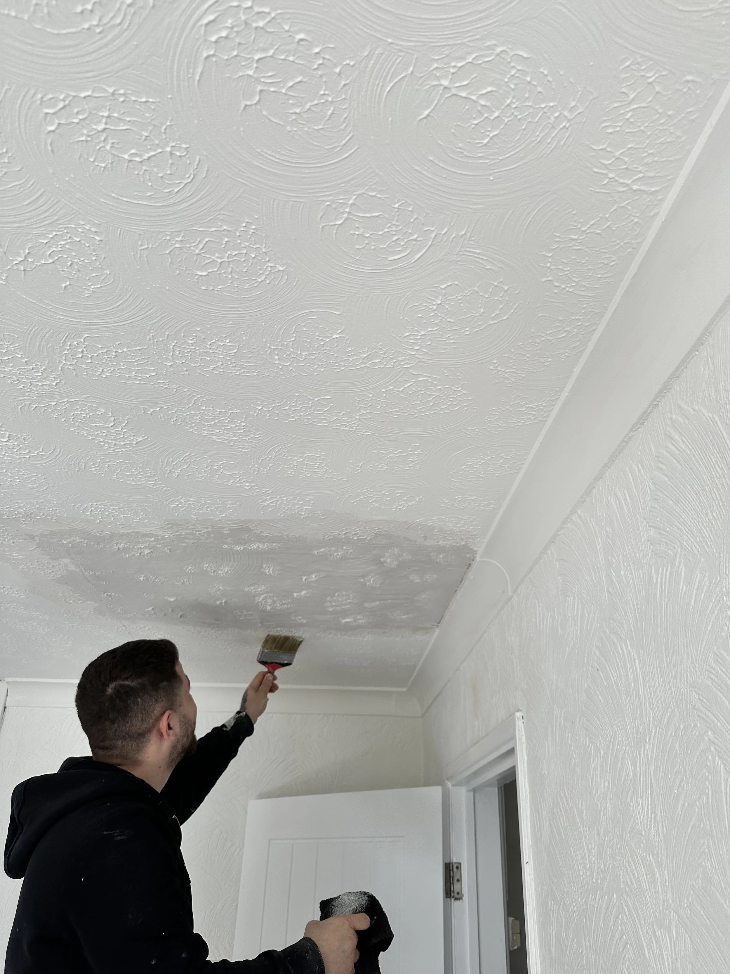 A man painting a textured ceiling white with a paintbrush while holding a paint roller in his other hand.
