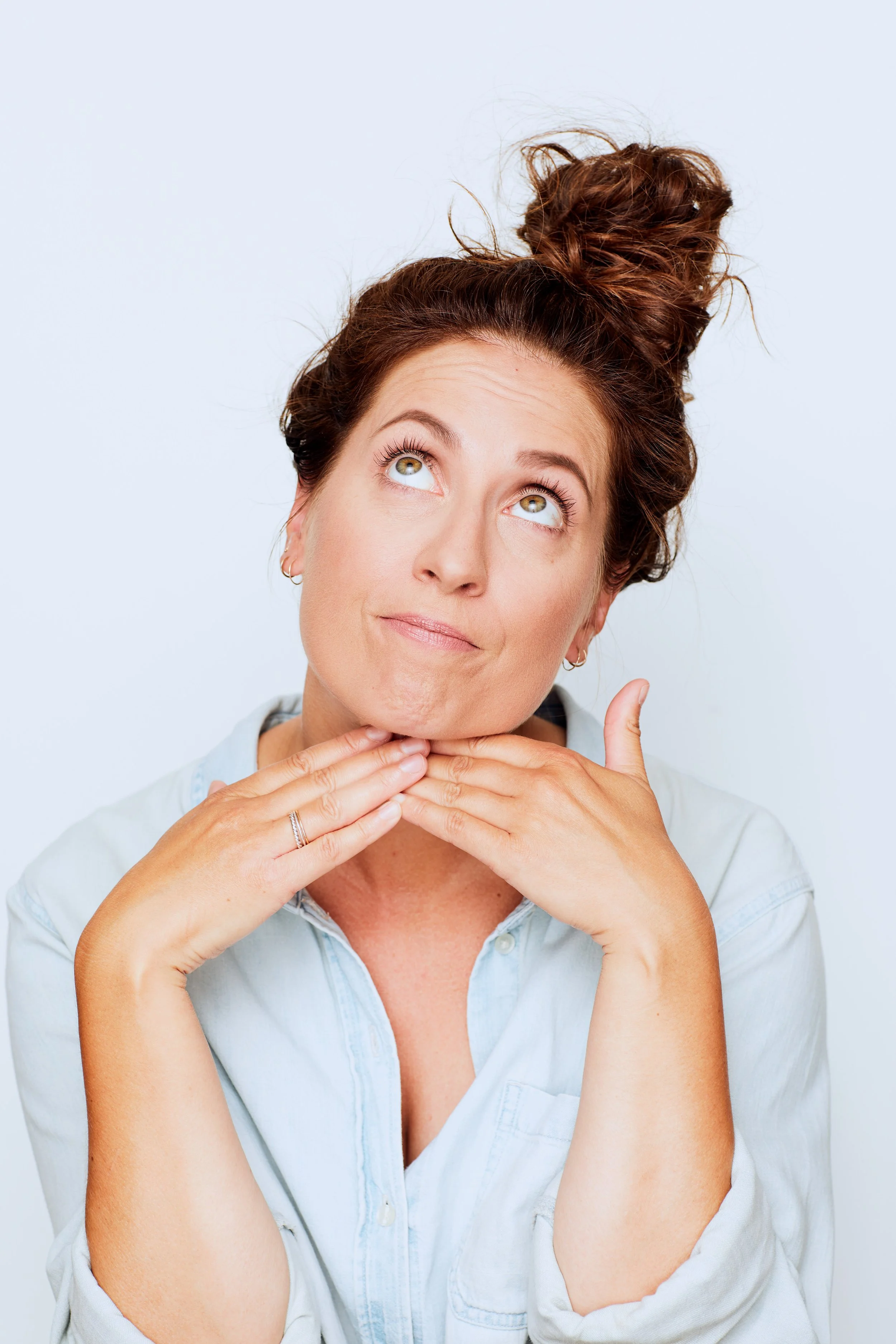 Woman with curly brown hair, styled in a messy bun, holding her chin with her hands, looking up thoughtfully, against a plain white background.
